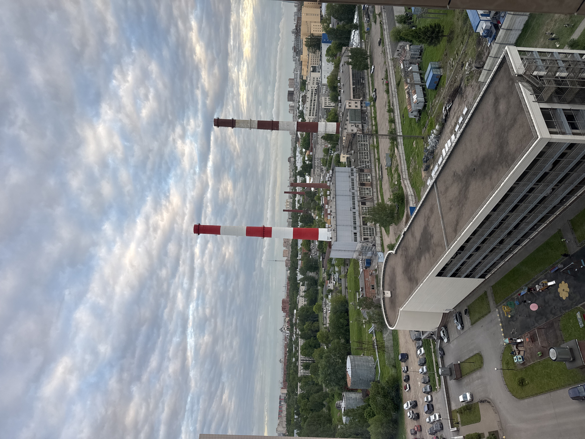 The image is a high-angle view of an urban landscape, likely taken from a tall building. The scene includes industrial structures with tall red and white striped smokestacks dominating the left side of the frame against a cloudy sky. A mix of residential and commercial buildings is visible in the distance, interspersed with greenery suggesting parks or tree-lined streets. Closer to the viewpoint, there are buildings with flat roofs, and parking areas with cars. A playground can be seen to the bottom right of the image. The general setting indicates a city in Russia.