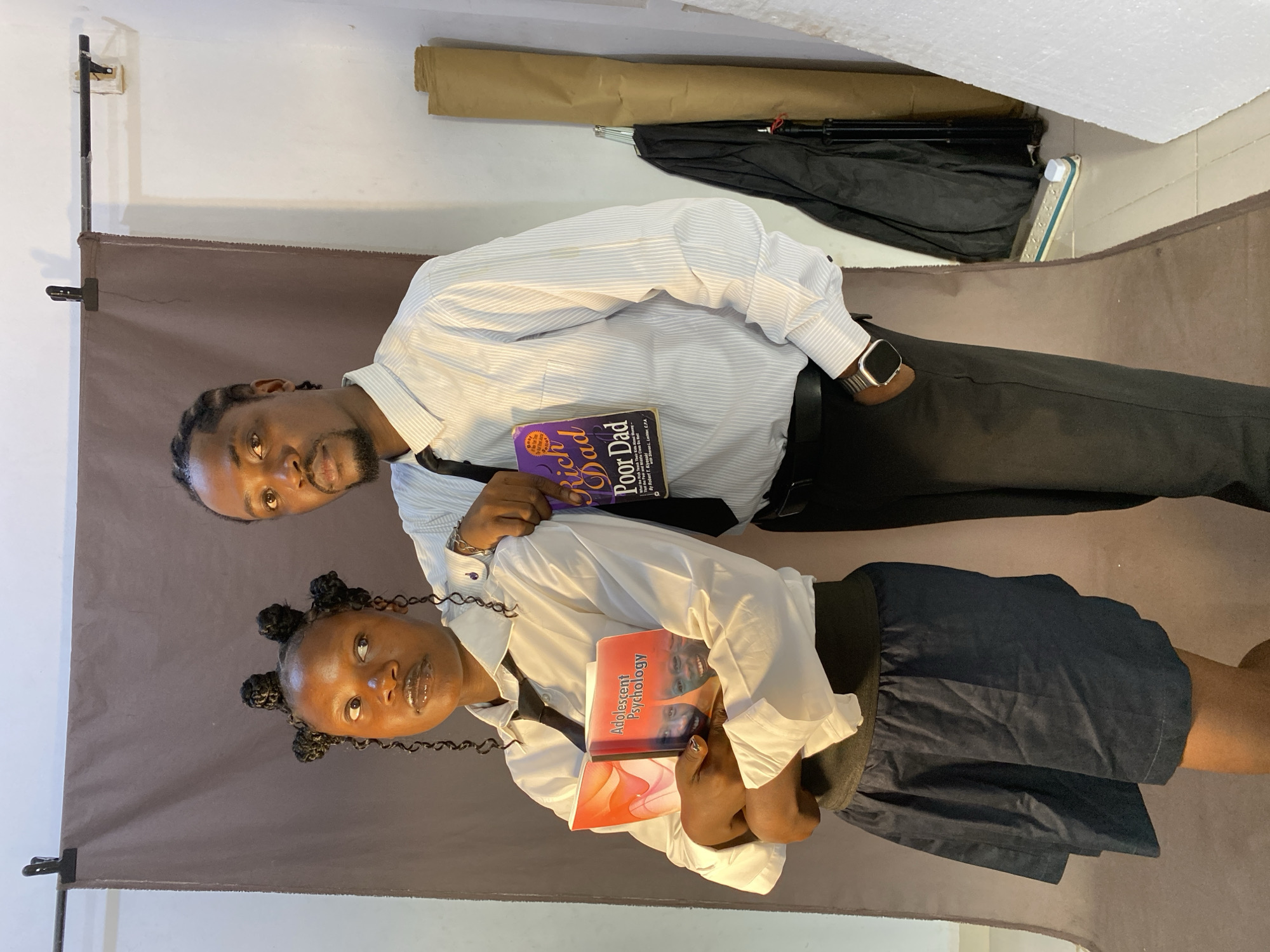 An adult male and a younger female are captured in a portrait-style photograph, standing in an indoor setting. The image is taken in Bukuru, Nigeria.
The man, positioned behind and slightly to the right of the female, has dark skin, braided hair, and a beard. He wears a light-colored, striped button-up shirt, a dark tie, dark trousers, and a silver-toned smartwatch on his left wrist. He holds a purple book titled "Rich Dad Poor Dad" by Robert T. Kiyosaki in his left hand.
The female, in the foreground, also has dark skin and her hair styled in small buns with braided extensions. She wears a white button-up shirt, a dark tie, and a dark skirt. She holds two books in her hands; the top book is titled "Adolescent Psychology," while the bottom one is orange with an abstract design.
Both individuals are looking directly towards the camera with neutral to slightly smiling expressions. They are posed in front of a brown fabric backdrop, which is secured by a metal stand and clamps. The surrounding white walls reveal photographic equipment such as a black umbrella, a rolled-up tan object (possibly another backdrop), and a white power strip, suggesting a makeshift photo studio setup. The books they hold are notable details, indicating themes of finance and education.