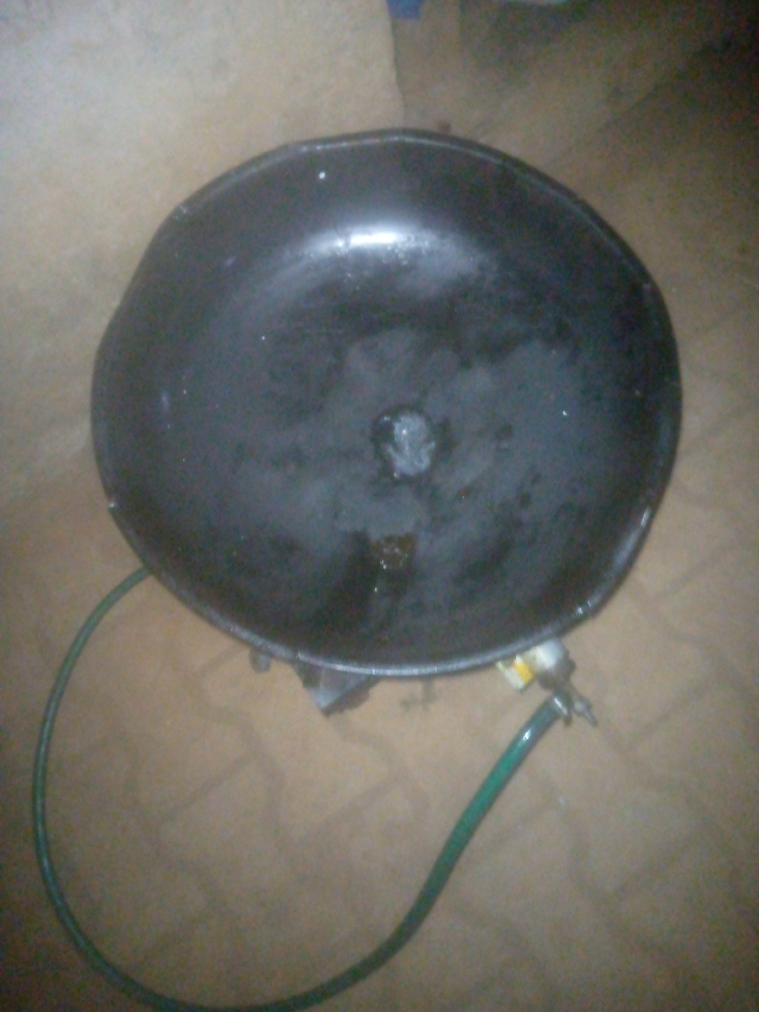 The image shows a black, bowl-shaped object with a green hose connected to its side. The interior of the bowl appears to have some liquid residue. The object is resting on a surface that is a light brown color. The location and further context are not available.