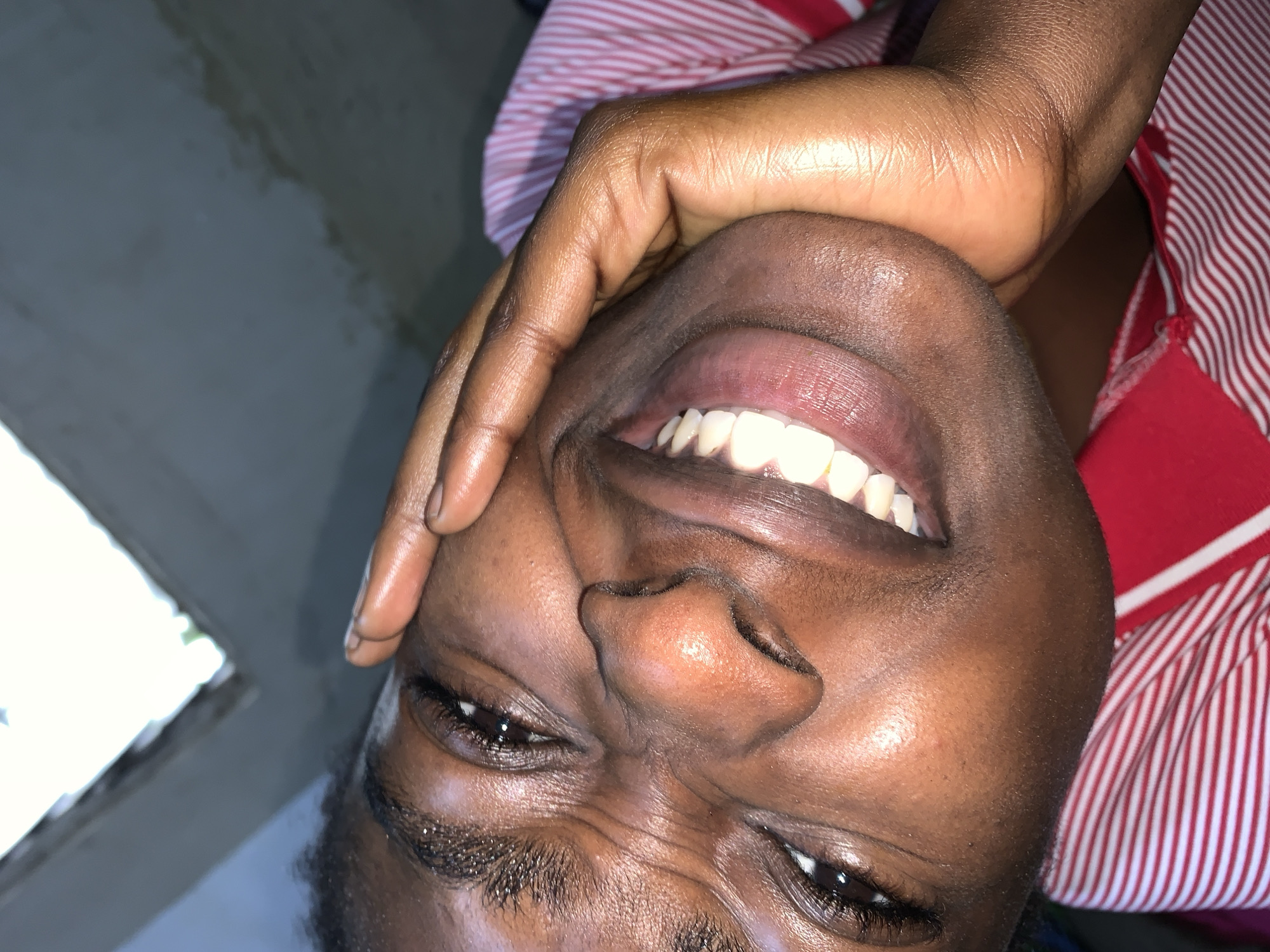 In a close-up, slightly angled shot, a person with dark skin is captured with their face turned towards the camera, one hand pressed against their cheek. Their eyes are partially closed and crinkled, suggesting a smile or laughter, and their teeth are visible in a wide grin. The person is wearing a red and white striped shirt. The background is blurred, but appears to show a gray wall with a bright opening at the top, possibly a window or skylight, hinting at an indoor or sheltered environment. The scene does not contain any information or visual cues that allow for the identification of a specific location within Rafin Par, Nigeria.