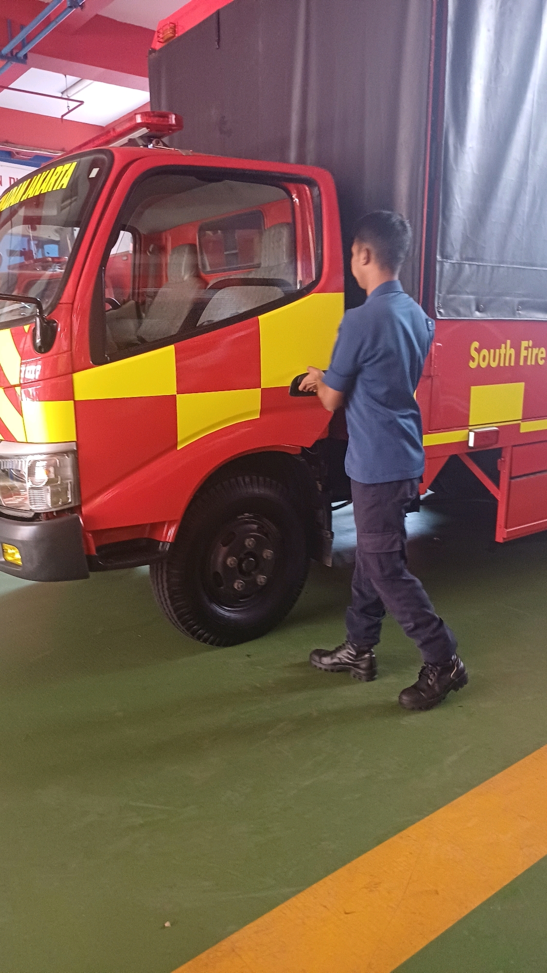 A side-view, medium shot captures a person wearing a dark blue uniform and dark trousers, standing next to a red and yellow checkered fire truck. The individual is facing the truck, with their right arm extended towards the door handle, appearing to be opening it. The fire truck, with the text "South Fire" visible on its side, is parked indoors on a green floor with a yellow stripe. The background is a dark gray tarp and red ceiling structure with lights. The scene suggests an operational context for the fire services within the city of South Jakarta, Indonesia.