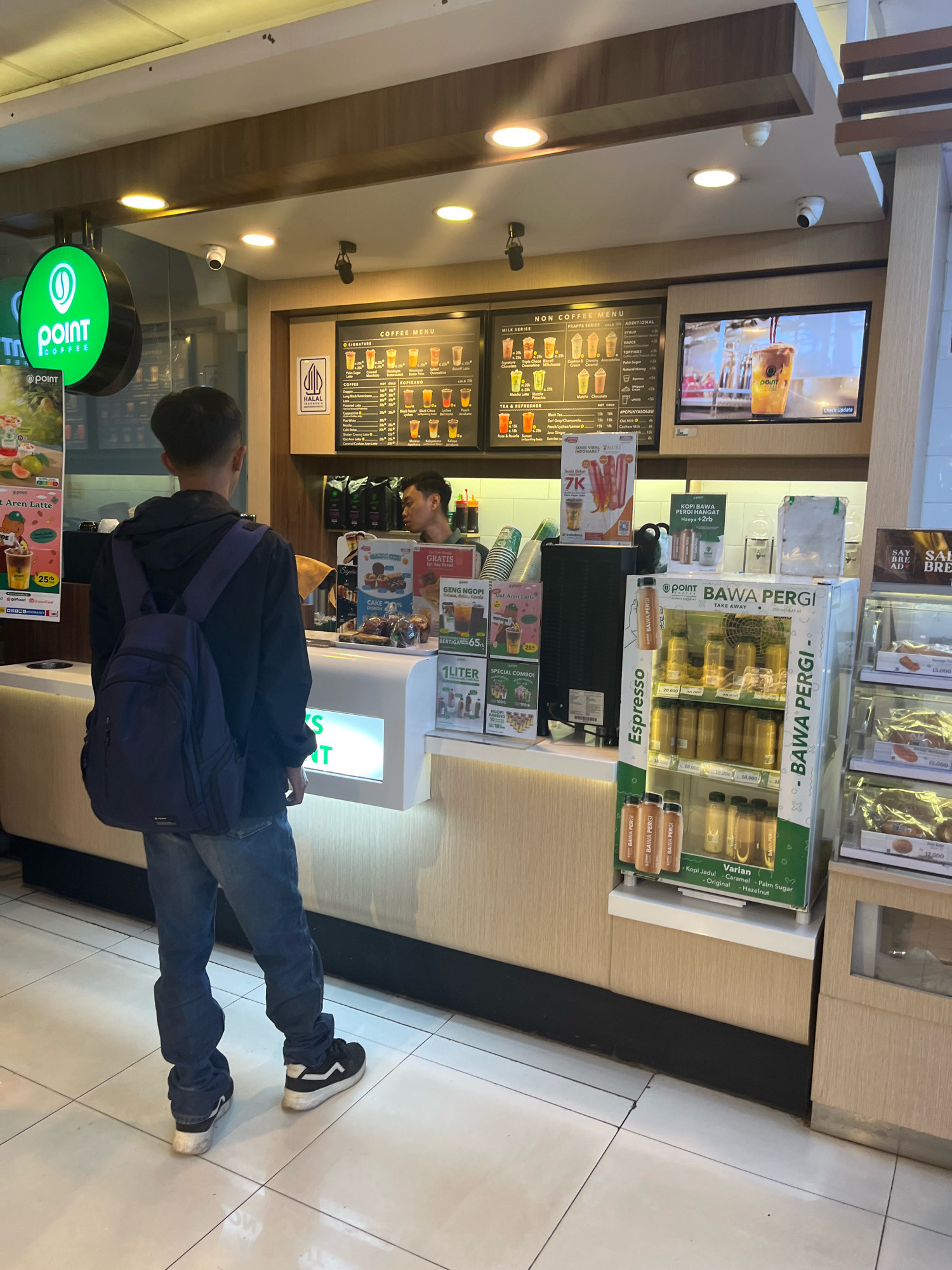 This image depicts a Point Coffee shop in Banguntapan, Indonesia. A young man wearing a dark hooded jacket, blue jeans, and a navy blue backpack stands facing the counter, waiting to order. He has short dark hair and is looking towards the menu board. Behind the counter, a male barista with short dark hair and a light-colored shirt is visible, interacting with the customer. The coffee shop environment is evident, with a menu board displaying various coffee and non-coffee drinks, a refrigerated display case filled with bottled beverages, and shelves stocked with products. Promotional posters and signage are also present. The flooring is tiled, and the counter area appears clean and well-lit. The overall scene is characteristic of a modern coffee shop setting.