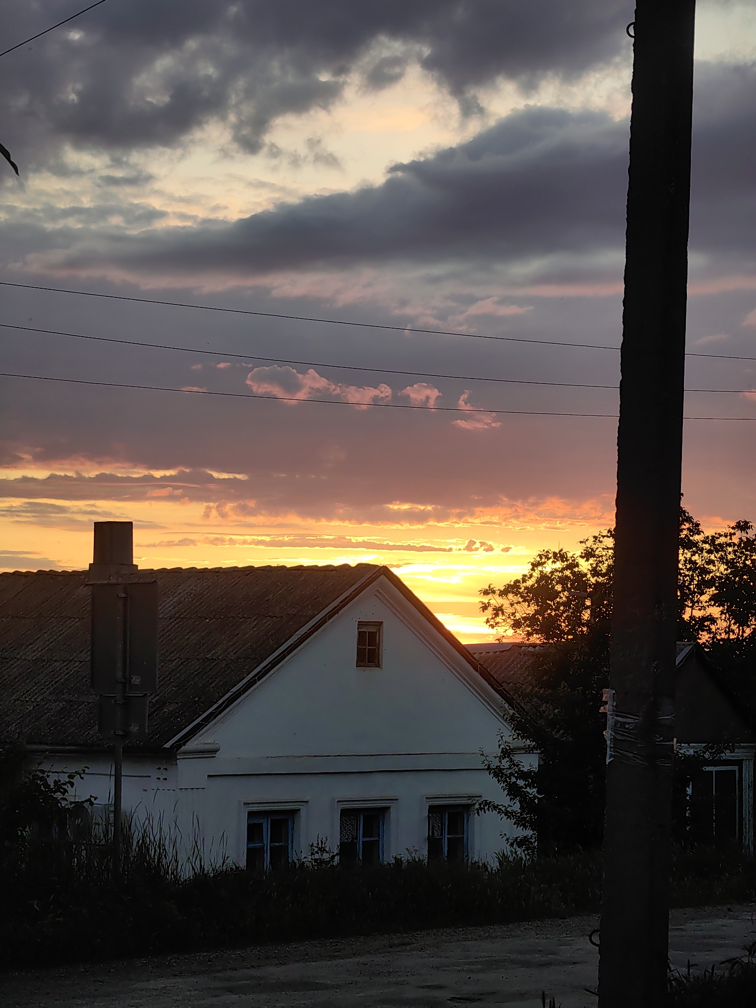 A rural scene unfolds at sunset in Zuyanske rural settlement, Ukraine. The sky is a dramatic canvas of orange, yellow, and pink clouds, transitioning to darker grey and purple hues towards the top. A silhouetted utility pole dominates the right side of the frame, its rough texture visible against the vibrant sky. In the mid-ground, a white house with a dark, tiled roof is partially visible, its gable end featuring a small, dark window. A chimney stands prominently on the left side of the roof. Tall, dark grass and foliage obscure the lower part of the house and the surrounding ground. To the right of the house, dark tree silhouettes are visible against the glowing sunset, their branches stark against the colored sky. Power lines are strung across the upper portion of the image, adding a geometric element to the natural scene. The overall impression is one of a quiet, rural evening as the day concludes.