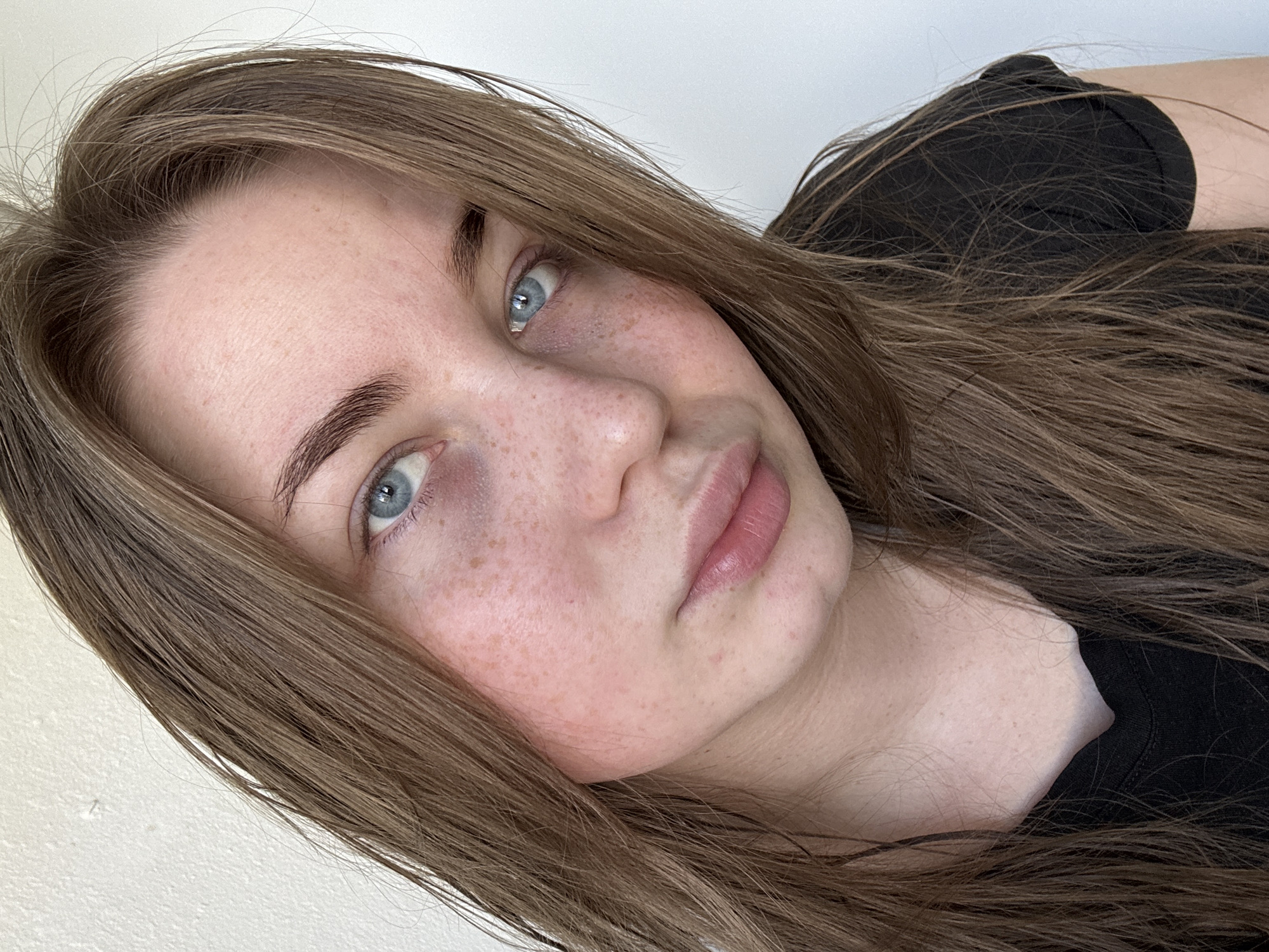 A close-up, slightly angled selfie of a fair-skinned young woman with light brown hair and blue eyes. Her face is freckled, and her lips are a natural pink. She is wearing a black top. The background is a plain white wall. The photograph was taken indoors. No information available indicates the location in the city of Chaikovsky, Russia.