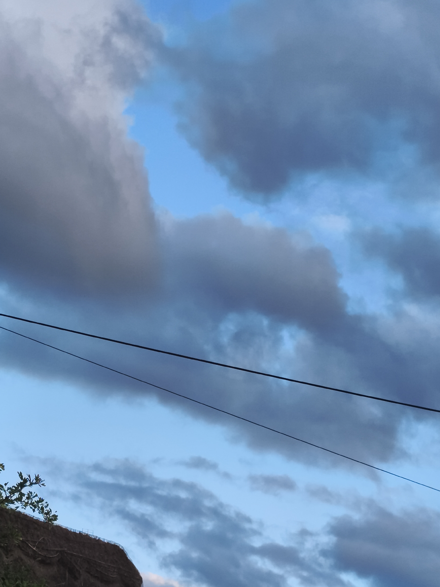 The image presents an outdoor scene dominated by a sky filled with a mix of dark, heavy greyish-blue clouds and clear patches of light blue sky. Large, dense cloud formations stretch across the upper and middle sections of the frame, with distinct areas of bright blue sky peeking through, particularly in the top left and middle right.
Two dark power lines traverse horizontally across the mid-ground, with the upper line appearing thicker and more defined, while the lower one is thinner and runs slightly below it, both descending gently from left to right.
In the bottom left corner, a portion of a brown, textured embankment or hillside is visible, possibly covered in soil or a protective netting. Green foliage and tree branches extend into the frame from the top of this embankment.
The lighting suggests either early morning or late afternoon, characterized by a soft, cool illumination. The presence of both dark clouds and clear sky indicates a dynamic weather condition, possibly on the cusp of changing. No people, animals, or visible text are present in the image. The scene is a tranquil yet atmospherically dramatic capture of the sky and its immediate surroundings.