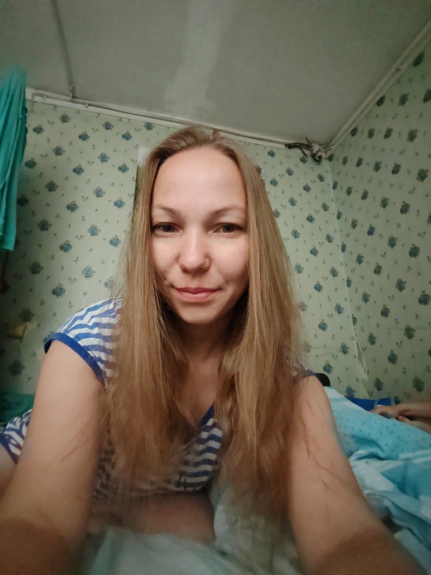 A medium shot selfie taken indoors shows a fair-skinned woman with long, light brown hair and blue eyes. She is wearing a blue and white striped t-shirt. The woman is positioned in the center of the frame, looking directly at the camera with a slight smile. Her arms are resting on a light blue blanket. The background consists of a light green wall with a subtle floral pattern and a white ceiling with visible molding. To the left of the woman, a teal garment hangs. The overall setting appears to be a bedroom.