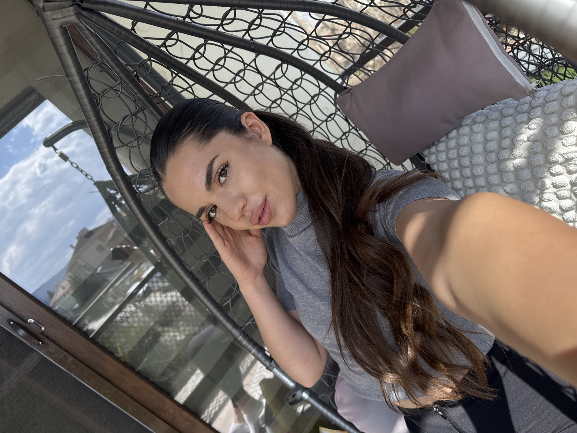 A female subject, appearing to be an adult, is depicted from a low-angle perspective, likely a selfie. She has dark brown hair pulled back in a slick ponytail and wears a grey, ribbed, mock-neck top and dark bottoms. Her left hand is positioned near her ear/temple. She is seated in a dark, woven, egg-shaped hanging chair, which contains a white textured cushion with a bobble pattern and a rectangular light purple cushion with a white zipper.
The setting appears to be a balcony or outdoor area. To the left, a dark brown wooden window or door frame is visible. The background, largely seen through a glass reflection, shows a bright blue sky with scattered white clouds and the upper portion of a light-colored residential building with a reddish-brown tiled roof. Sparse tree branches are also reflected. Above the subject's head, part of the dark metal suspension chain and hook for the hanging chair is visible against the sky. The architecture of the reflected building is consistent with residential areas found in Sofia, Bulgaria. The scene is illuminated by strong natural sunlight, casting shadows.