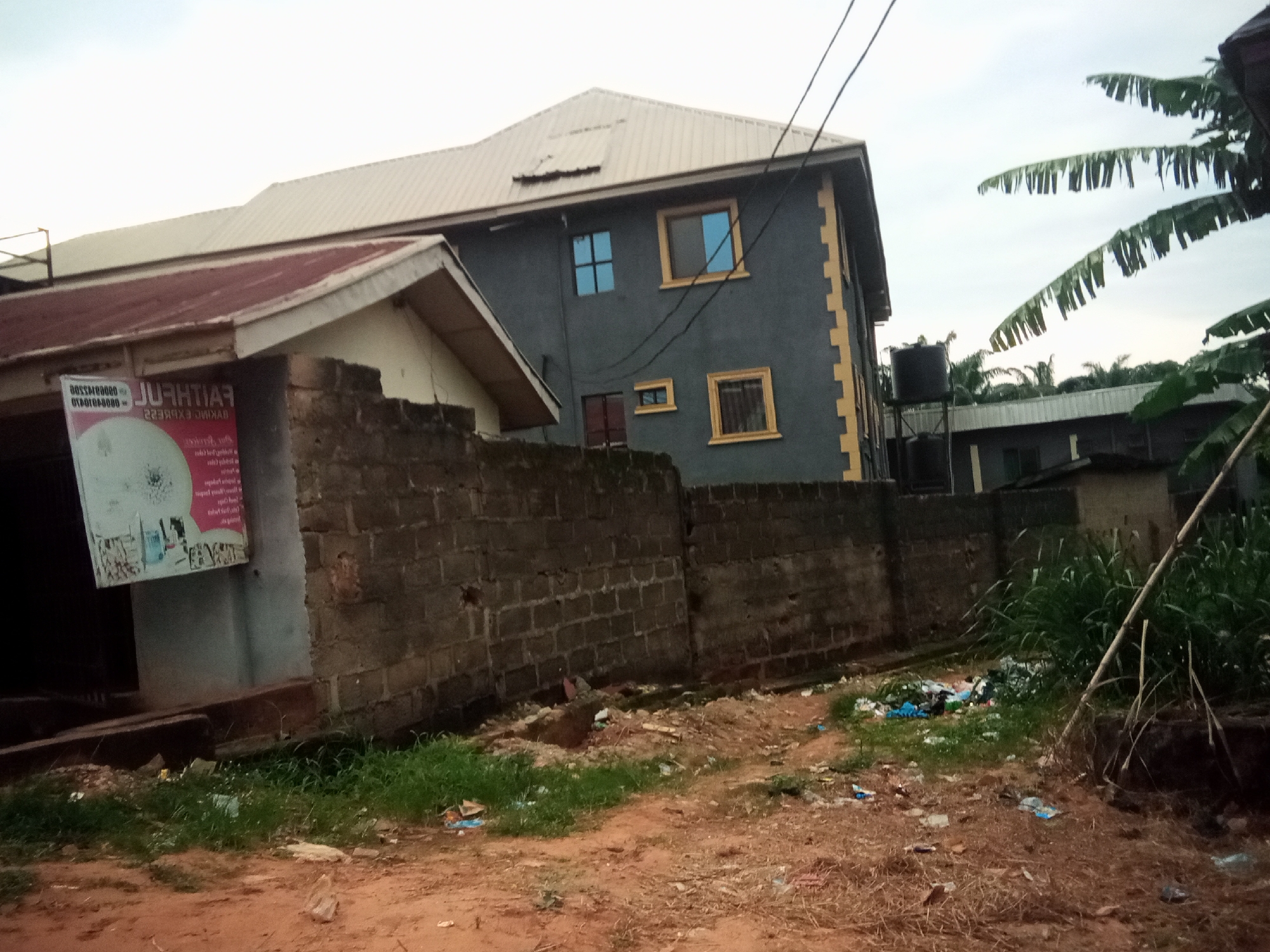 The image depicts a scene in Oko Town, Nigeria, featuring several buildings and a brick wall. The main building in the foreground is a two-story structure with a grey facade and yellow decorative accents. It has several windows, some with blue tinted glass, and a corrugated metal roof. To the left, another building with a red corrugated metal roof is partially visible, featuring a sign with text in what appears to be a Latin script. A black water tank is mounted on a metal stand behind the grey building.
A brick wall runs across the middle ground of the image, separating the buildings from a dirt and grass area. This area is littered with various debris, including plastic bottles and bags, suggesting an unkempt environment. In the background, more buildings and palm trees are visible under an overcast sky. The ground in the foreground is reddish-brown dirt. There are no people visible in the image.
