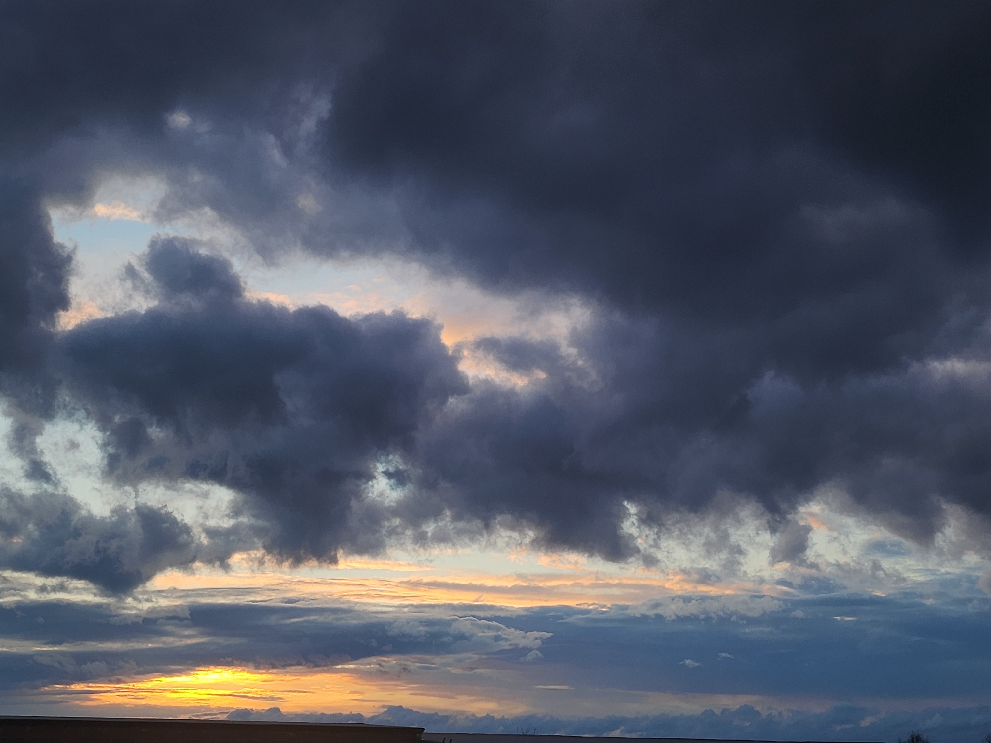 The image displays a dramatic and expansive sky filled with numerous clouds, captured during either sunrise or sunset over Krasnogorskoye, Russia. The upper and central portions of the frame are dominated by heavy, dark gray and deep blue clouds, appearing thick and voluminous, suggesting an overcast or potentially stormy condition.
Through and between these darker cloud masses, lighter patches of pale blue sky and softer, white to light gray clouds are visible, allowing hints of ambient light to break through. Towards the lower third of the image, a vivid display of warm colors emerges, indicative of the sun's low position. A bright, intense yellow and orange glow is prominent at the bottom left, casting a radiant light that illuminates the undersides of the clouds with golden and peachy hues. This creates a striking contrast with the cooler, darker tones above.
A narrow, dark silhouette forms the very bottom edge of the image, representing the horizon line, likely a distant landscape or ground feature. The overall scene emphasizes the raw power and beauty of the sky, with the interplay of dark clouds and brilliant light creating a dynamic and awe-inspiring atmosphere. There are no people, specific man-made objects, or visible text in the image.