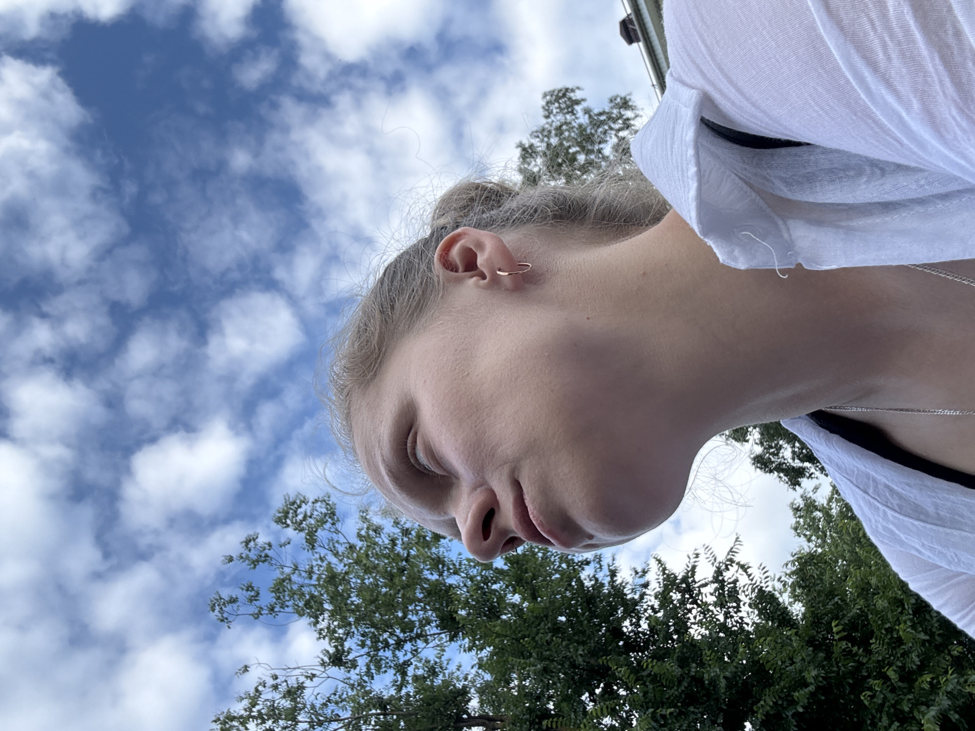 The image is a close-up, low-angle shot of a young Caucasian female looking up into a cloudy blue sky. She has fair skin, blonde hair pulled back in a bun, and is wearing a simple gold hoop earring in her left ear. Her eyes are closed, and her mouth is slightly open as if in thought or contemplation. She is wearing a white, textured collared shirt. Green trees are visible in the background, and a portion of a building with a dark roof is peeking out from the right side of the frame. The lighting suggests it is daytime, likely outdoors in a natural setting. The image appears to have been taken in Mikhaylovsk, Russia.