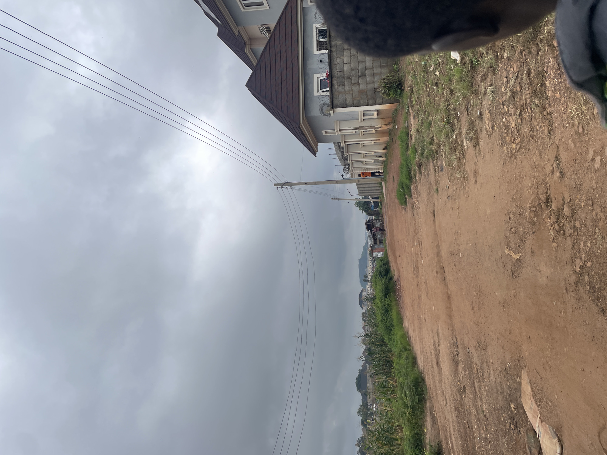 The image depicts a dirt road in a suburban area of Abuja, Nigeria, under a cloudy sky. On the right side of the road, a modern two-story house with light blue walls and a brown roof is visible, surrounded by a grey concrete block wall topped with barbed wire. A person in an orange shirt stands near the gate of this property. Further down the road, on the left, is a field of tall green crops, possibly maize, with more houses and vegetation visible in the distance. Utility poles with multiple overhead wires are present along the road. The road itself is unpaved and appears to be in a state of disrepair, with visible potholes and uneven surfaces. The foreground shows the shoulder of the road with sparse grass and some debris. The overall impression is one of developing infrastructure in a rural or semi-rural setting within Abuja.