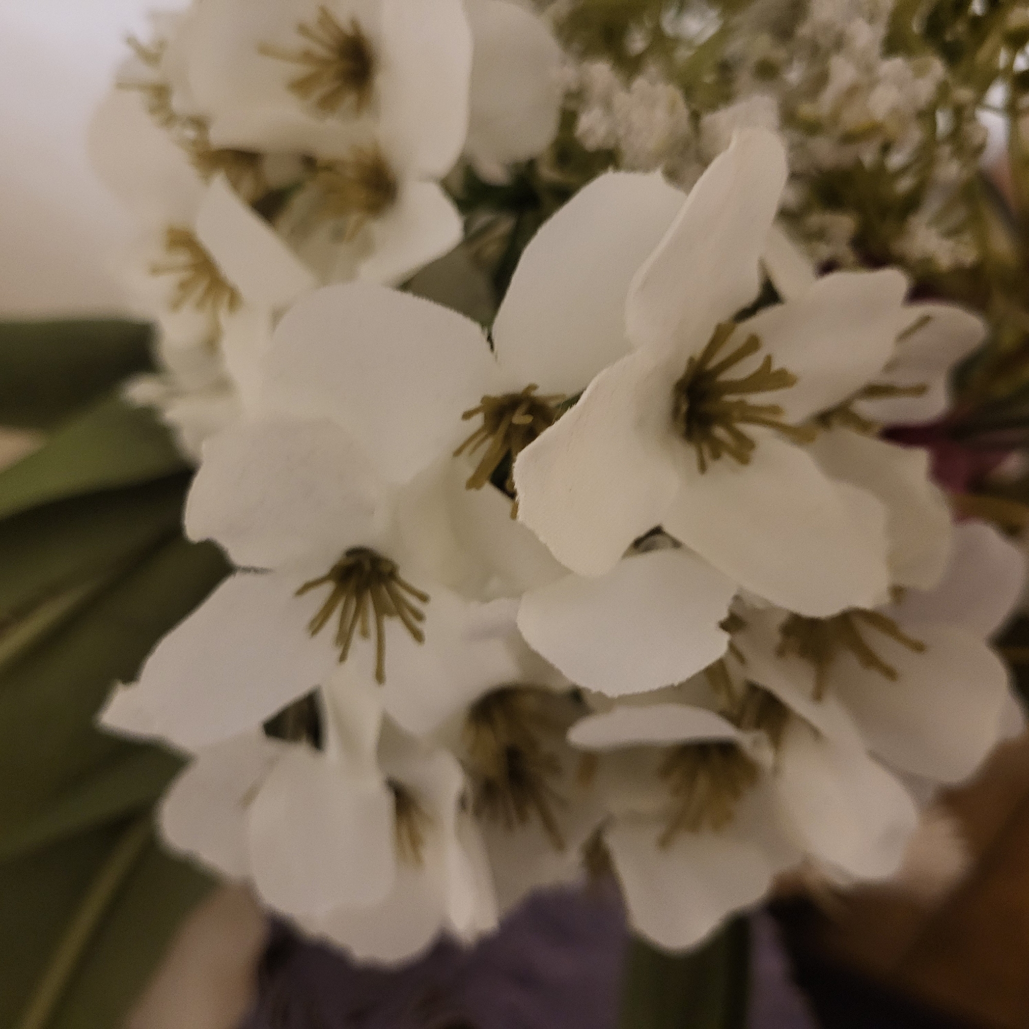 The image shows a close-up view of a bouquet of artificial white flowers with yellow-brown centers. Some green leaves are visible in the background, along with smaller white flowers resembling baby's breath. The overall focus is soft, and the lighting appears to be indoor and muted. The location is not discernible from the visual information provided; there are no landmarks or features to indicate that the image was taken in Yerevan, Armenia.