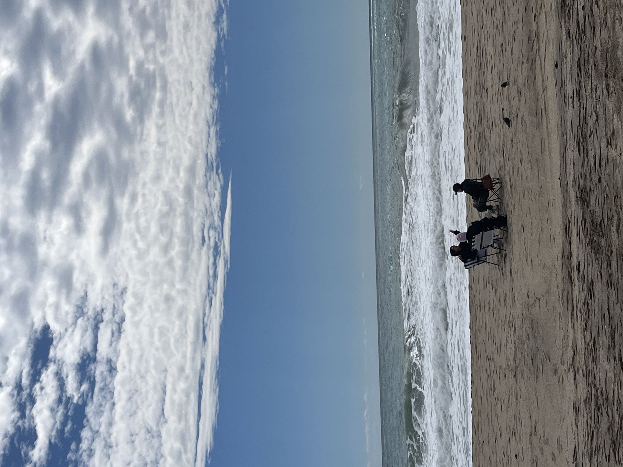 A beach scene is depicted in the image. The sky is divided between a large cloud formation on the left and a clear blue section. The ocean occupies the central area, with breaking waves near the shore. Two individuals are sitting in chairs near the water's edge on the sandy beach. The location is in Alanya, Turkey.