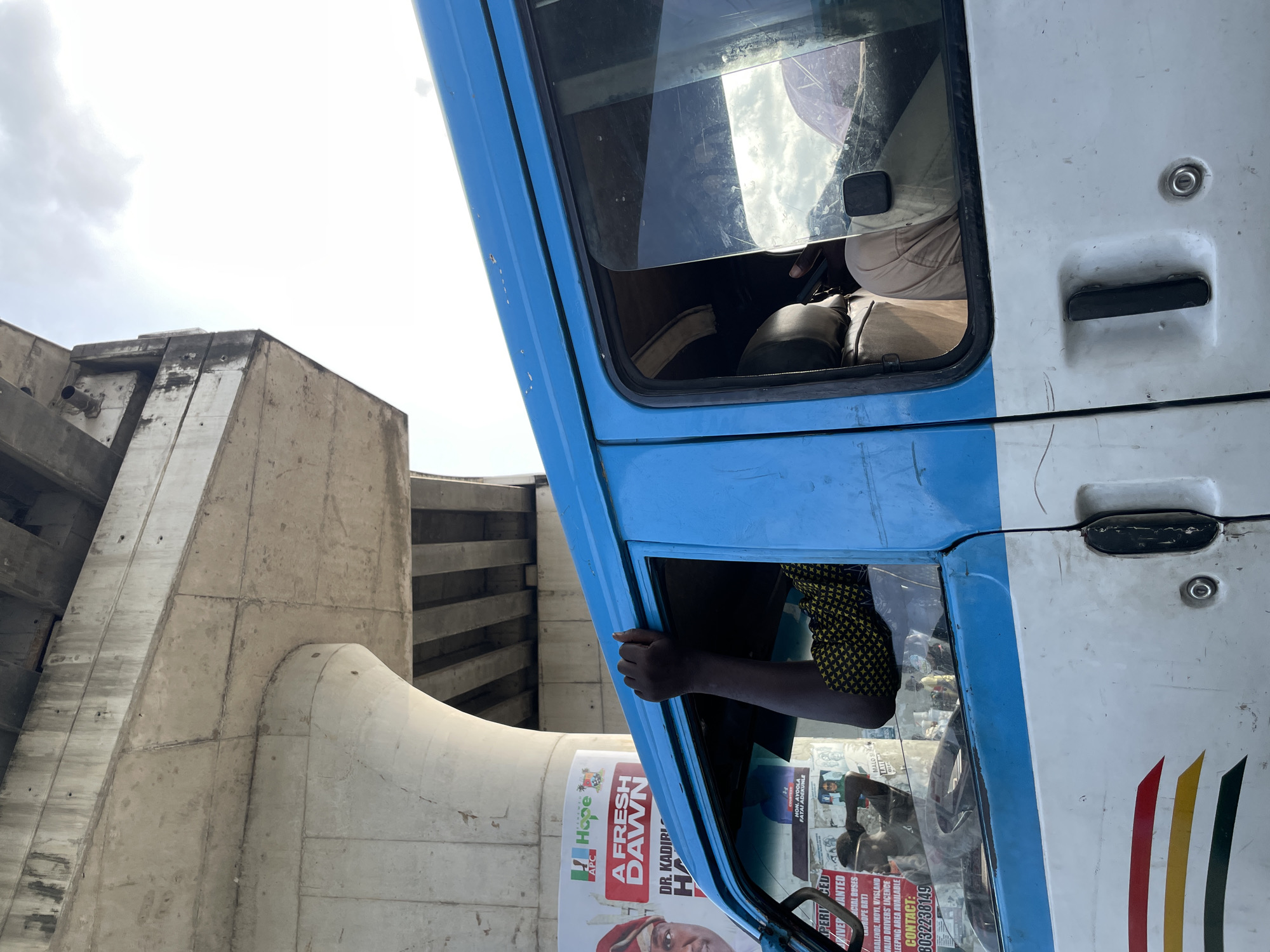 An outdoor, medium shot captures a blue and white van on a street in Lekki, Nigeria, under a large concrete overpass. A person's arm, wearing a yellow and black patterned shirt, is visible extending from the open driver's side window, hand resting on the frame. The van's exterior shows signs of wear, with faded paint and some scratches. Posters are affixed to the interior window, one featuring a smiling person and text that reads "A FRESH DAWN." The sky is partly cloudy. The overpass is constructed of weathered concrete with visible support structures and a rough underside.