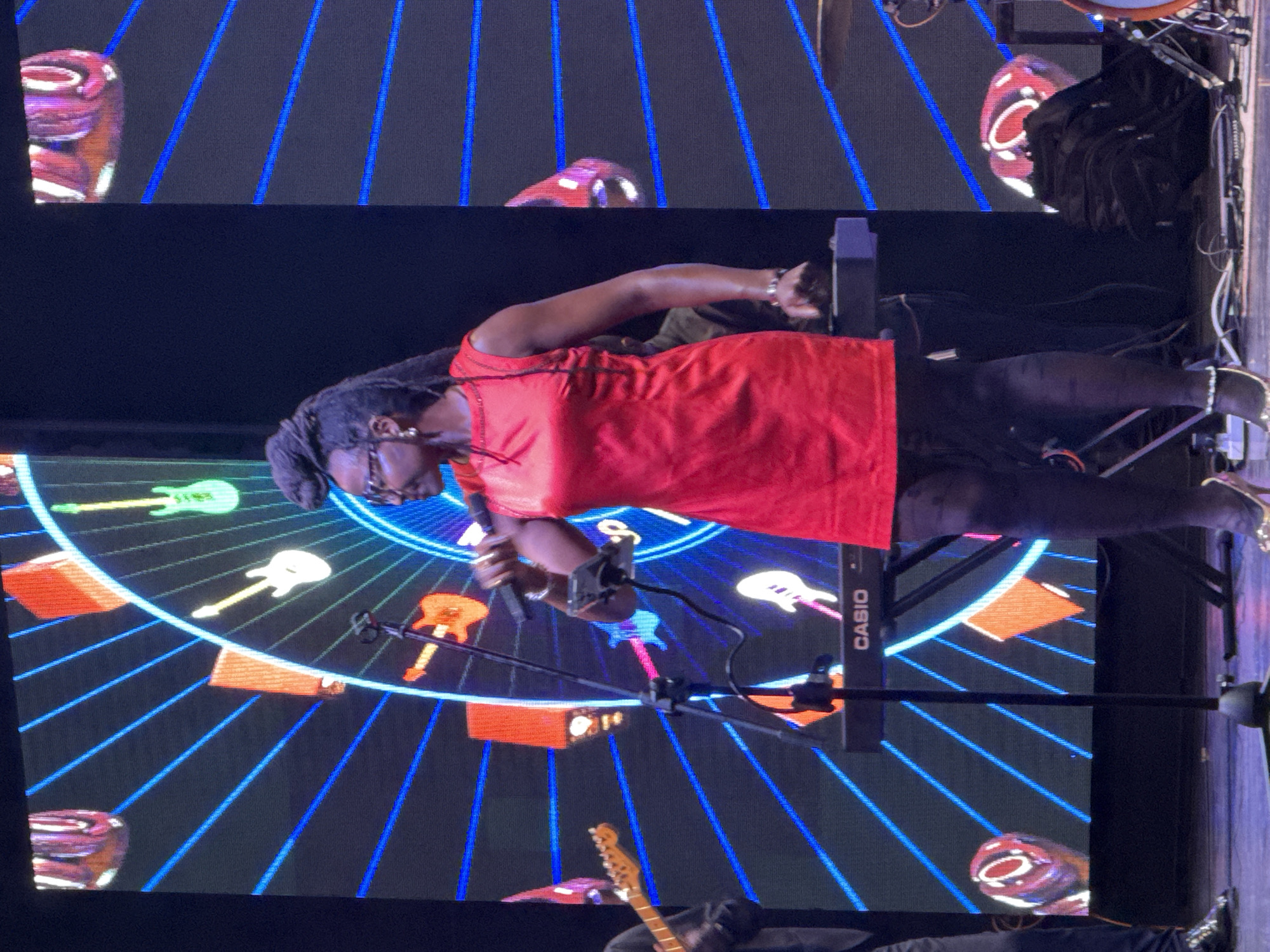 A Black woman wearing glasses and a red sleeveless dress is singing into a microphone on a stage. She has her hair in a bun. She is wearing black patterned tights and gold high-heeled shoes. A keyboard with "CASIO" written on it is behind her. In the background, a digital screen displays a circular graphic with blue lines emanating from the center, featuring images of guitars and orange diamond shapes. The woman is in the foreground, with parts of other musicians visible to her left and right. The setting appears to be an indoor performance venue. The image was taken in Manavgat, Turkey.