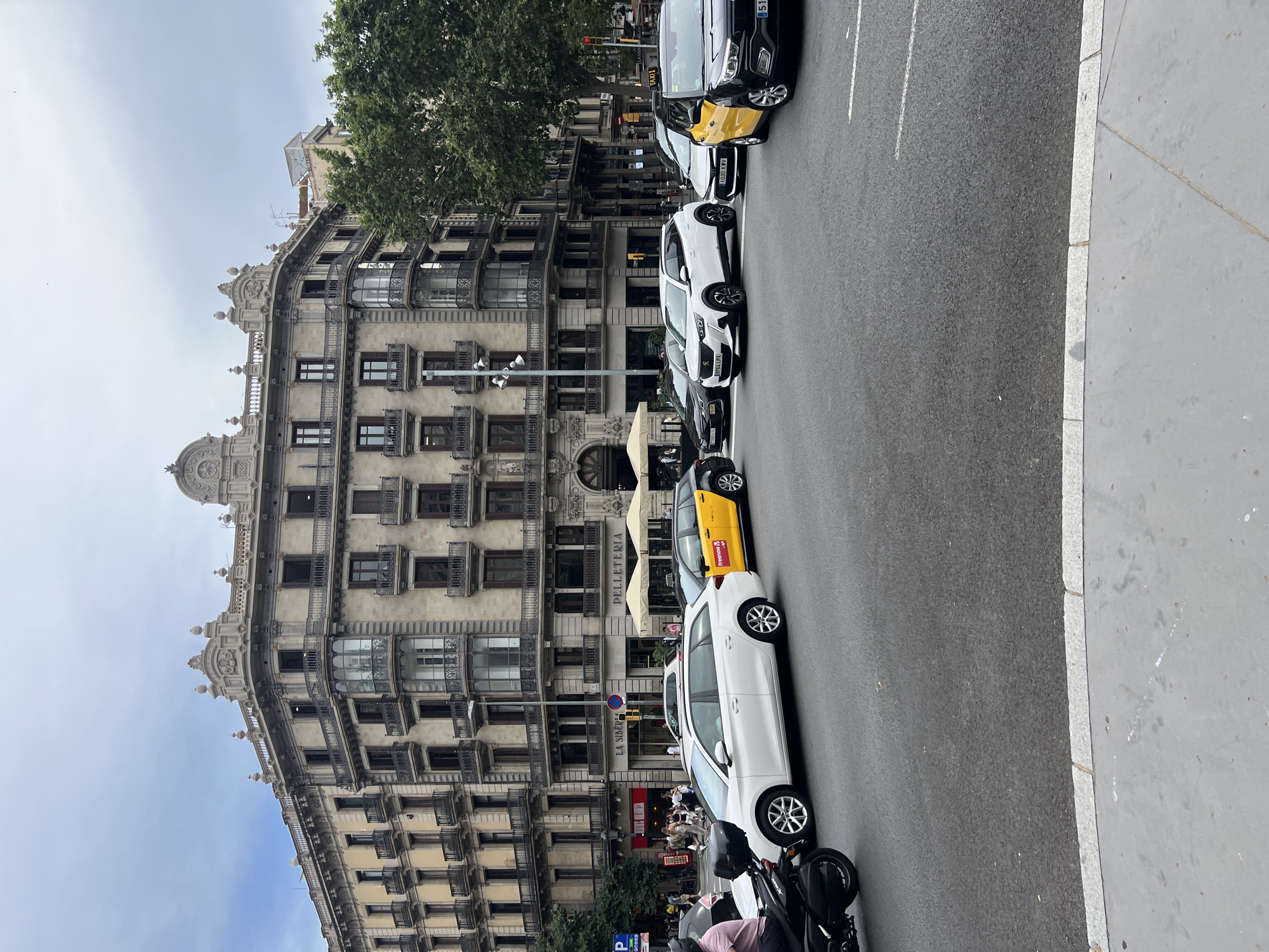 This image depicts a street scene in Barcelona, Spain, likely in a dense urban area with historic architecture. In the foreground, a grey concrete sidewalk runs along the edge of a paved road. The road is occupied by several vehicles, including a black motorcycle on the far left, a white station wagon, a black and yellow taxi with "freenow" branding, a white SUV, another white car, and a black SUV that appears to be a taxi. The vehicles are positioned as if in traffic.
Behind the vehicles, a multi-story building with ornate architectural details dominates the background. The building features numerous balconies with wrought-iron railings, arched windows, and decorative cornices. Signs for "LA SIMBORA" and "PELLETERIA" are visible on the ground floor. Green trees are visible to the right of the building. The sky is overcast with a few birds visible in the upper left corner. The overall impression is a bustling city intersection under a cloudy sky.