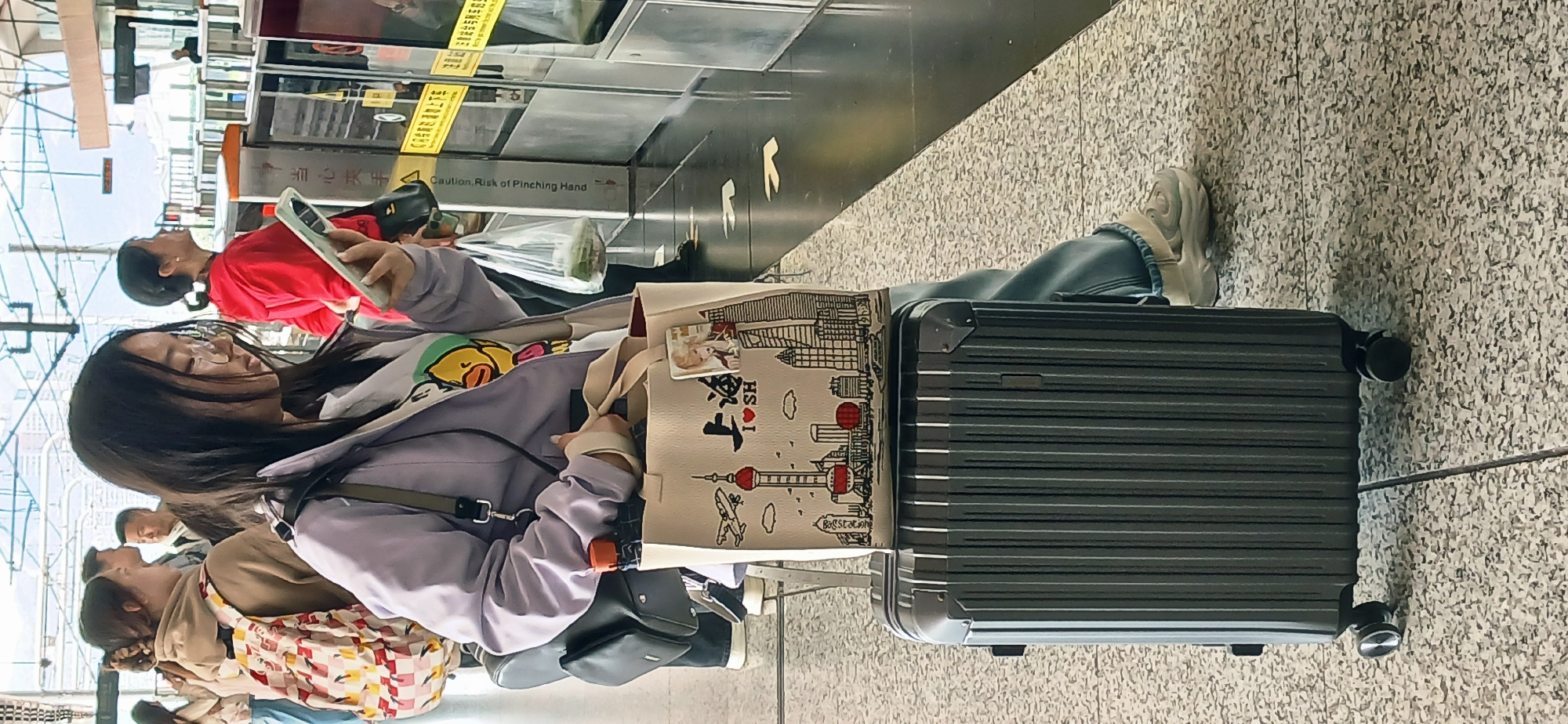 The image captures a scene at an elevated railway station in Chongqing High-tech Zone, China. In the foreground, a young woman with long black hair is seated, her attention directed at a smartphone she holds in her hands. She is wearing a light purple jacket over a white t-shirt with a cartoon graphic, blue jeans, and white sneakers. A shoulder bag rests beside her. A large black suitcase is positioned to her right, and a shopping bag with illustrations of Shanghai landmarks sits on top of it. Several other people, including a woman in a red shirt also holding a smartphone, are visible in the background, waiting near an automated ticket barrier. The station platform has a gray speckled floor and features black automated gates with warning labels. Overhead structures are visible, suggesting the platform is elevated.