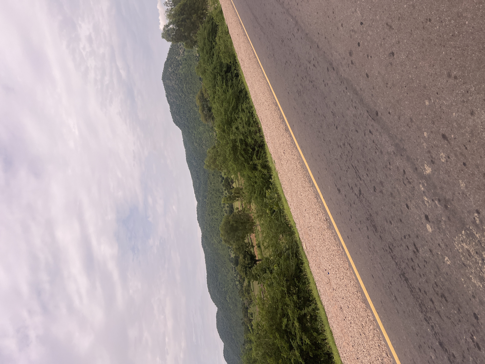 This media file shows a scenic view of a road running alongside a lush green mountainside in Minda, Nigeria. The road, paved with asphalt, has a distinct yellow line marking its edge, separating it from a gravel shoulder. The mountains, covered in dense vegetation, dominate the left side of the image, with their slopes stretching into the distance. A band of green trees and bushes runs along the base of the mountains, bordering the road. The sky above is overcast with light, wispy clouds, suggesting a diffused, possibly humid, atmosphere typical of this region. The scene appears to be captured during daylight hours, though the specific time of day is difficult to ascertain due to the cloudy conditions. No people or specific objects are prominently featured; the focus is on the natural landscape and the man-made road. The overall impression is one of a tranquil, rural setting, possibly a view from a moving vehicle.