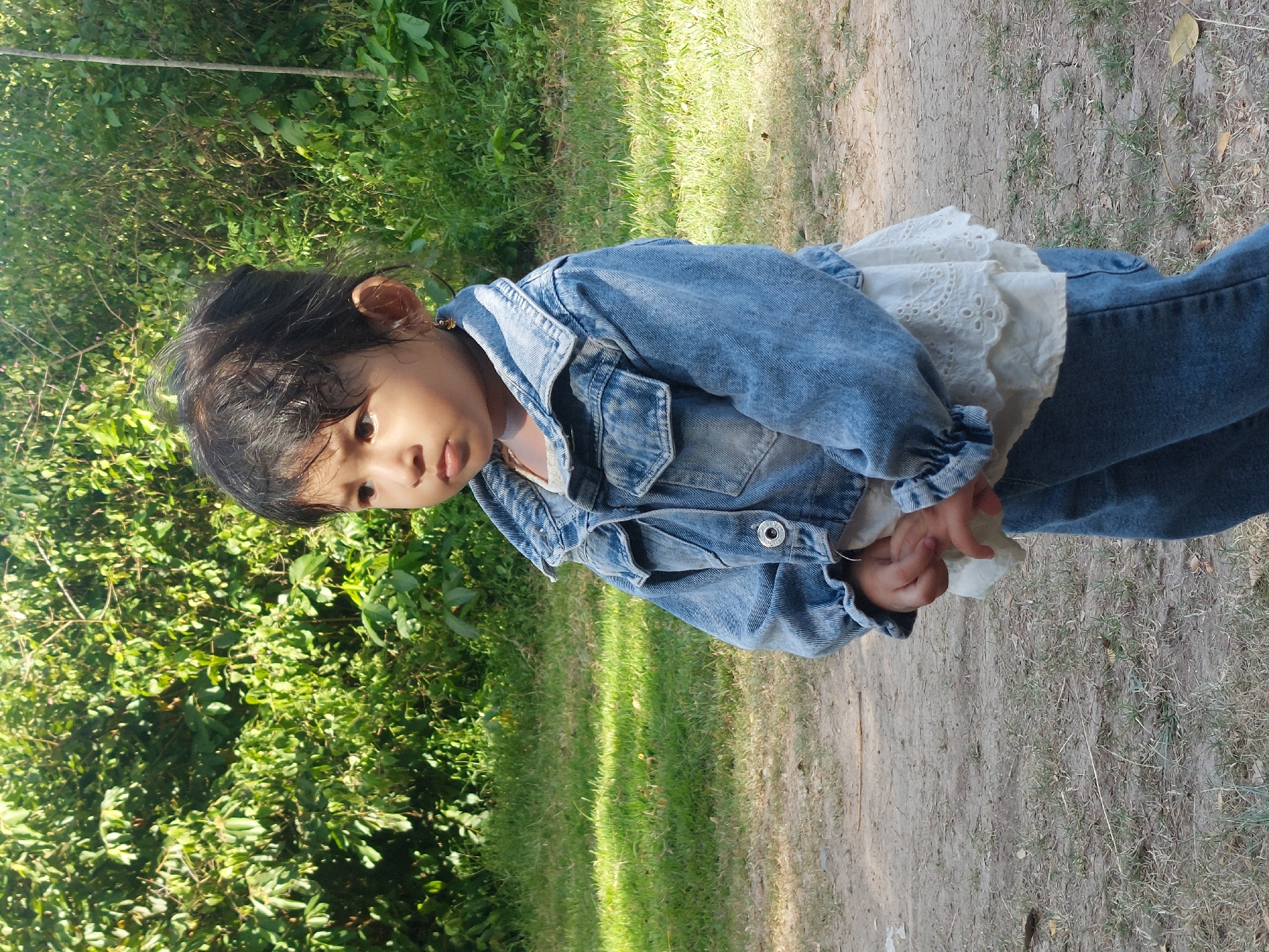 A young child stands on a dirt path next to green foliage. The child wears a denim jacket and jeans over a white, lace-trimmed top. The location appears to be in a natural environment. The scene is taken during daylight hours. There is no verifiable evidence in the image to confirm its location in Jamuan, Indonesia.