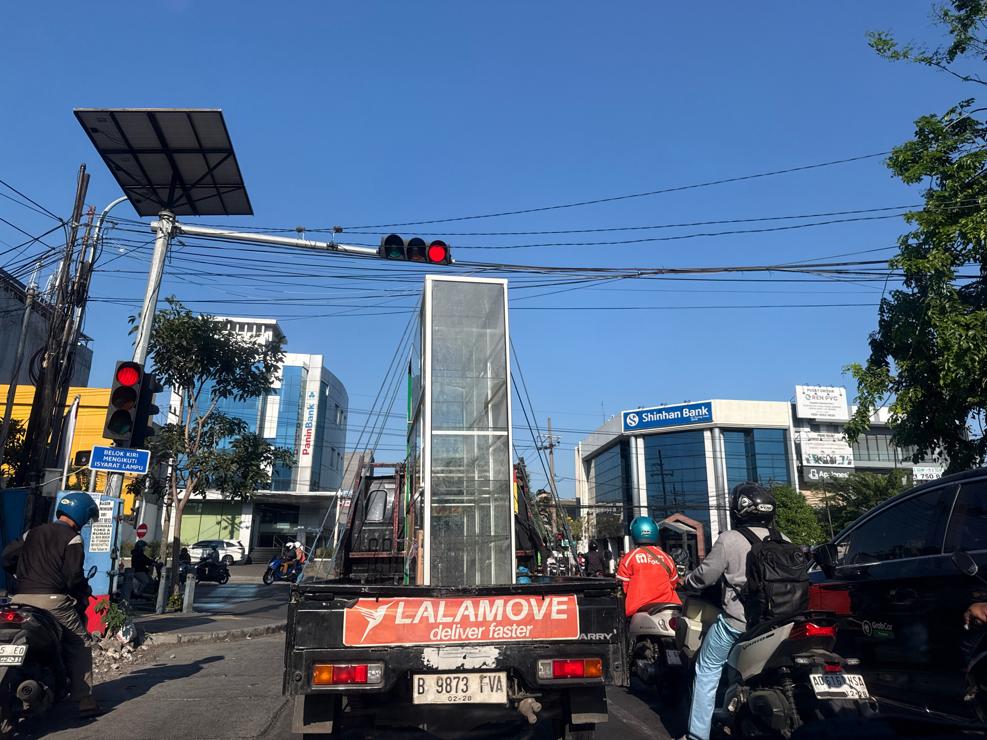 A busy street scene in Surabaya, Indonesia, is captured under a clear blue sky. The foreground is dominated by the rear of a black pickup truck adorned with a bright orange "LALAMOVE deliver faster" logo. The truck's license plate reads "B 9873 FVA" and shows an expiration date of "02-28".
Several motorcycles are present. A person wearing a blue helmet and a dark jacket is seated on a motorcycle to the left. To the right, a rider in an orange shirt and teal helmet, likely a Lalamove delivery person, is on a white motorcycle. Another motorcycle with a rider in a grey jacket and black helmet, carrying a backpack, is in the center right.
The background reveals a typical urban cityscape. Tall buildings line the street, including "PaninBank" and "Shinhan Bank," identifiable by their signage. A traffic light is visible, displaying a red signal. Electrical wires crisscross overhead, suspended from poles. Trees with green foliage add a touch of nature to the concrete environment. The overall impression is one of daily urban activity, with traffic and commerce in full swing.