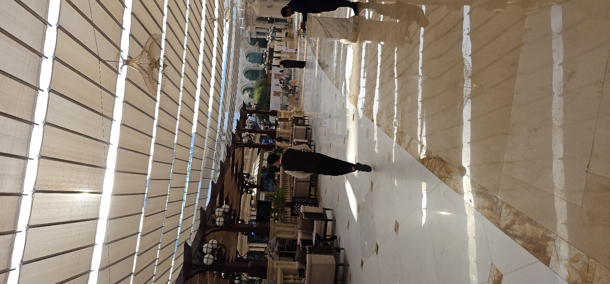 The image depicts an interior view of a large, modern building with a high, canopied ceiling and a polished marble floor. The ceiling consists of a grid of beige fabric panels supported by a metallic structure, allowing sunlight to filter through, creating a pattern of light and shadow on the floor. Several ornate chandeliers are suspended from the ceiling.
In the foreground, a man wearing a white shirt and a dark vest is walking away from the camera, his back to the viewer. He is on a white and light brown tiled floor. Further into the background, several people are visible. On the right side, a man in a dark suit is standing near a marble pillar, appearing to interact with something on the pillar. In the center background, there is a seating area with upholstered chairs and tables, and more people are seated and standing. The architecture features ornate columns and arched structures, suggesting a luxurious or public space, possibly a hotel lobby, a shopping mall, or a similar venue in Doha, Qatar. The overall impression is one of spaciousness and natural light.