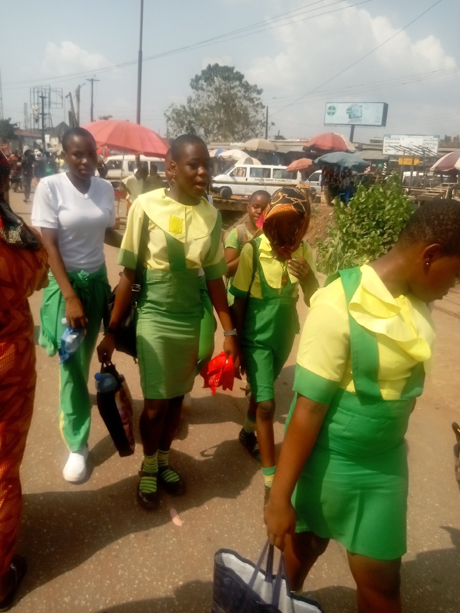 The image displays a daytime street scene in Onitsha, Nigeria, under bright, sunlit conditions with some cloud cover. Six young females are in the foreground and mid-ground, moving generally across a light-colored paved surface. Four of these individuals wear school uniforms consisting of green pinafore-style dresses or green trousers/leggings combined with yellow shirts or blouses. One student in the foreground wears a white t-shirt with green trousers and holds a blue water bottle. Another student holds a dark bag and a blue water bottle, while a third holds a piece of red fabric. A partially visible individual on the far left wears an orange patterned garment. Prominent shadows are cast on the ground, indicating direct sunlight.
In the background, a busy market environment unfolds, featuring numerous vendor stalls covered by various colored umbrellas, including red, white, and blue-green. Several white minibuses or vans are present on the street; one clearly displays text that includes "TRANSPORT COMPANY OF ANAMBRA STATE," confirming the regional location. Utility poles and multiple electrical lines crisscross the sky. A large billboard with a green logo is visible in the distance, alongside additional smaller signs advertising services such as "NEW REGISTRATION" and "CASH IN CASH OUT ELECTRICITY BILLS & DATA." A large tree and smaller shrubs are also visible within the urban landscape.