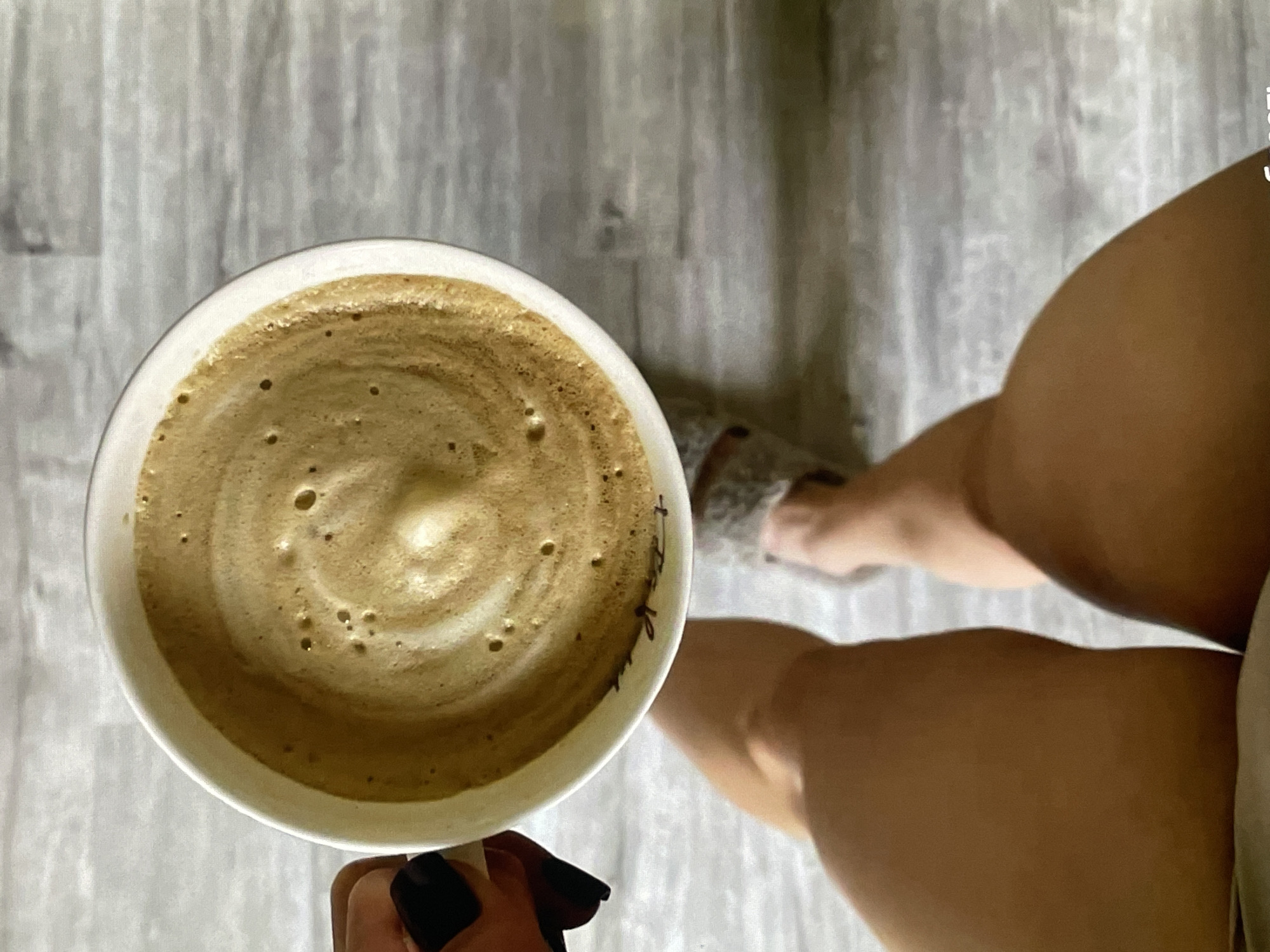 A top-down shot shows a white ceramic mug filled with a foamy coffee drink. The coffee has a swirl pattern on top. A hand with black nail polish is holding the mug. In the background, a person's legs are visible, wearing a fuzzy slipper on one foot. The scene appears to be set on a grey, wood-planked floor. There is no identifiable information or context to place this image in Neubrandenburg, Germany.