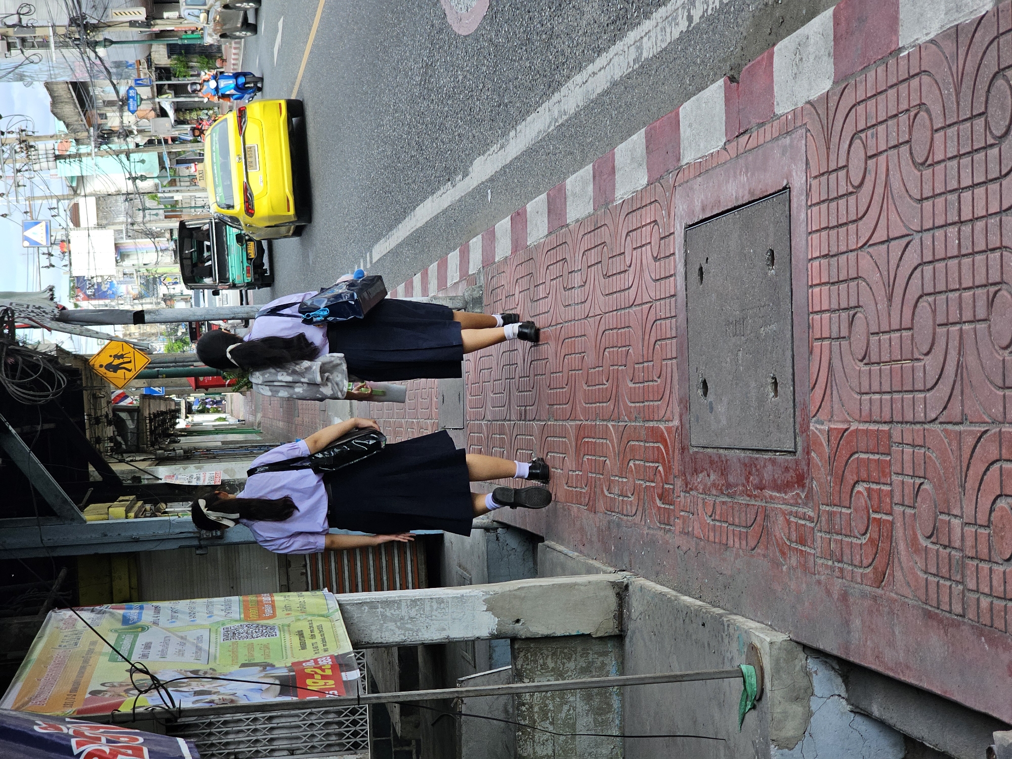 This image depicts a street scene in Bangkok, Thailand. Two young women in school uniforms are walking away from the camera on a red-tiled sidewalk. The woman on the left wears a light purple short-sleeved shirt and a dark pleated skirt, with her hair tied back in a ponytail. The woman on the right wears a patterned gray top and a dark skirt. Both are carrying bags.
The sidewalk is bordered by a red and white striped curb, with a street to the right. The street is paved and has visible traffic markings. Vehicles on the street include a yellow taxi, a green utility vehicle, and a blue scooter with a rider. In the background, there are buildings with visible overhead wiring, typical of urban areas in Thailand. A pedestrian crossing sign with two figures is visible on the left side of the street. Posters are displayed on a pole and a building facade on the left. The scene appears to be in an urban commercial or residential area.