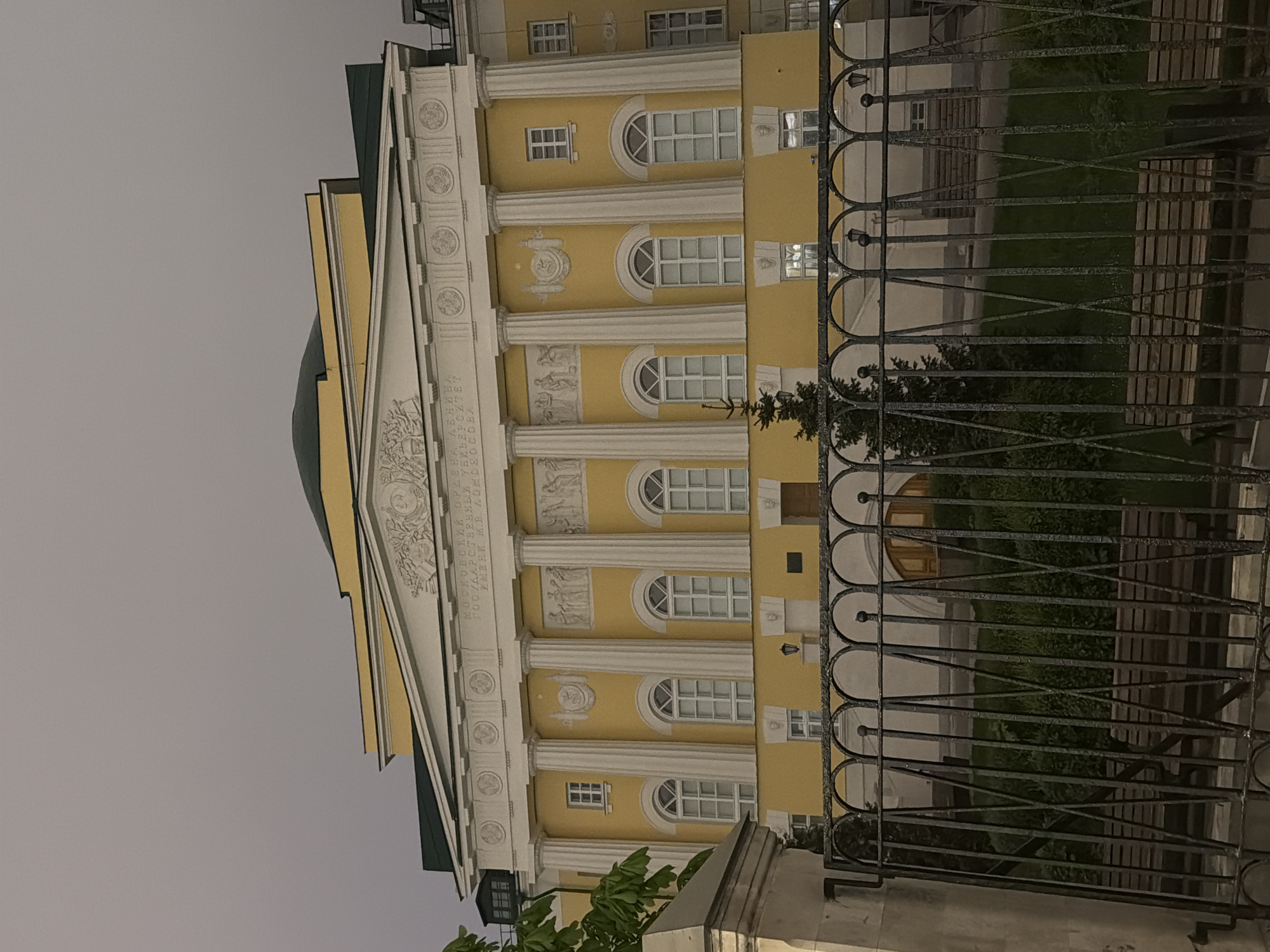 The image depicts the main building of Moscow State University in Moscow, Russia, seen from behind a wrought-iron fence. The building is a yellow structure with white columns and a green dome. It features arched windows and decorative elements above the columns. The sky is overcast.