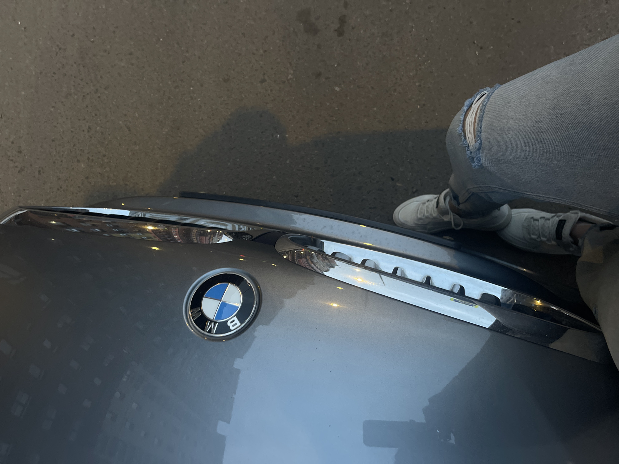 A BMW emblem is visible on the hood of a gray car. A person wearing ripped jeans and gray sneakers stands next to the car. The location appears to be an urban area, possibly in the city of Razvilka, Russia, although specific landmarks are not visible. The car's hood reflects the surrounding buildings.