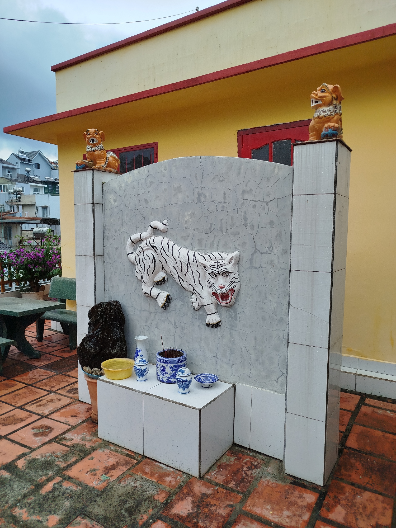 The image captures a courtyard or patio in Da Lat, Vietnam. The space is paved with terracotta tiles. Against a yellow building with red-trimmed windows sits a gray concrete altar. A white tiger sculpture is affixed to the central panel of the altar. The altar has two tiled columns on either side. A small white cabinet supports a collection of blue and white porcelain vases, incense sticks, and a yellow bowl. To the left, there is a stone formation, a green table, and a bench. In the background, other buildings are visible, indicating the location is in an urban area. Two lion statues sit atop the corners of the building that the altar sits in front of. The scene suggests a temple or spiritual place. The weather is overcast, which is typical of Da Lat. There are no people present in the image.
