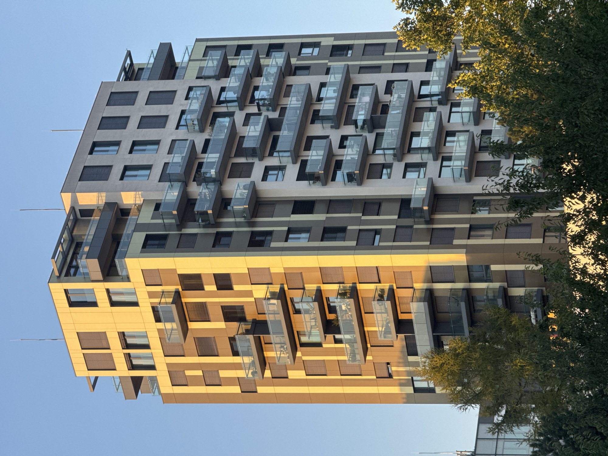 The image shows a modern, multi-story apartment building located in the city of Bratislava, Slovakia. The building has a distinctive facade with alternating panels of yellow and grey, with vertical brown stripes. Many of the apartments feature balconies with glass railings. The sunlight casts a warm glow on the yellow sections of the building. Trees with green foliage are visible in the foreground, partially obscuring the lower floors of the building. The sky is clear and blue.