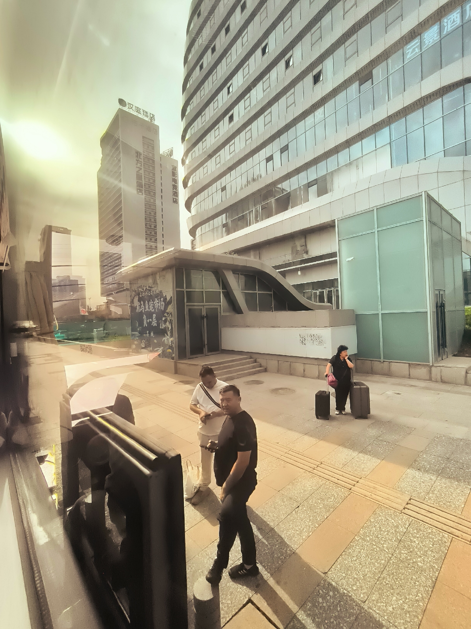 The image shows an exterior view of a modern building with glass facades and multiple stories, located in the city of Ürümqi, China. The sun is shining brightly, casting long shadows. In the foreground, a man in a black t-shirt and black pants, wearing black sneakers, walks along a paved sidewalk. He appears to be looking down. To his left, two other men are standing, one in a white t-shirt and the other partially visible in a black t-shirt. They are facing away from the camera. Further to the right, a woman wearing a dark outfit and a black handbag over her shoulder stands with two suitcases, one dark grey and the other black, both with wheels. She is looking to her right, her mouth slightly open as if speaking on a phone. The building has a contemporary design with clean lines and large windows. The sidewalk is made of rectangular paving stones with tactile paving in some areas. Other buildings, including a tall, slender tower with Chinese characters and signage, are visible in the background. The overall impression is of an urban environment, possibly near a transportation hub or commercial area, with people on the move with luggage.
