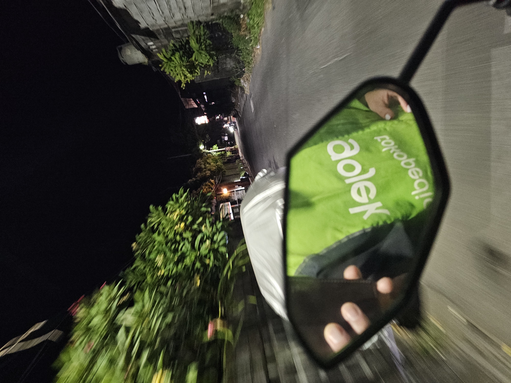 The image is a first-person perspective of someone riding a motorcycle at night on a narrow, paved road in Denpasar, Indonesia. The road is lined with buildings and dense foliage, and streetlights are visible in the distance. The rider's hand is visible in the foreground, holding onto the motorcycle's handlebars, and the motorcycle's side mirror reflects a bright green jacket with white lettering that appears to read "Gojek" in reverse. The motion blur indicates that the motorcycle is in motion, and the lighting suggests it is nighttime.