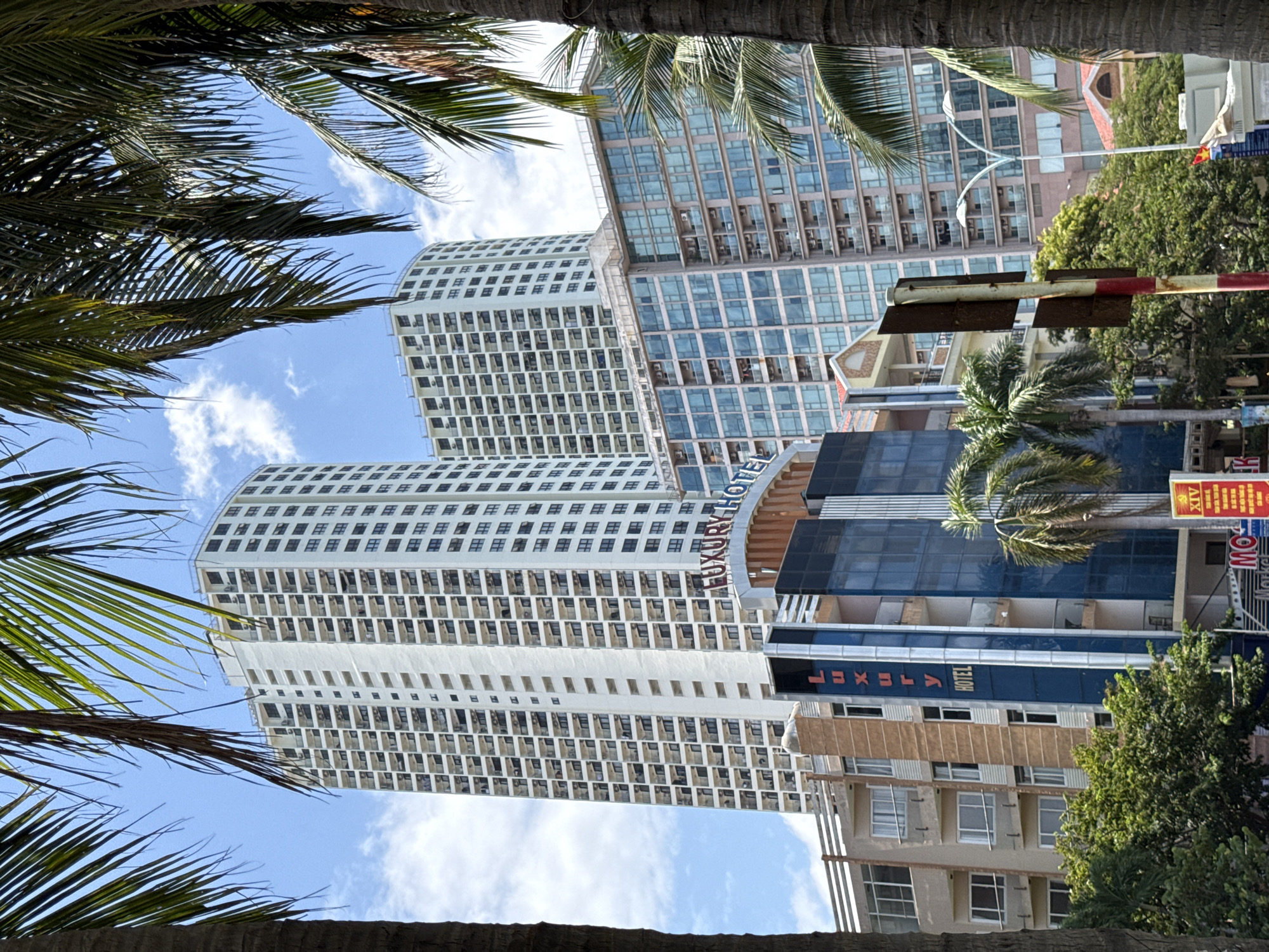 The image shows a street view in Nha Trang, Vietnam, with several high-rise buildings and palm trees. In the foreground, the fronds of two large palm trees partially obscure the view. The central focus is on a cluster of modern buildings. The tallest building is a white, multi-story residential tower with numerous balconies and dark-windowed units. To its right, another similar, slightly shorter white tower is visible. In front of these towers, a mid-rise building with a glass facade and multiple balconies is prominent, reflecting the sky. Below this, a building with a dark blue and glass exterior displays a vertical sign that reads "LUXURY HOTEL" in red lettering. To the left of the "LUXURY HOTEL" sign, a multi-story building with balconies and windows of varying sizes is seen. The street level shows more trees, a traffic light, and street signs. The sky is blue with scattered white clouds.