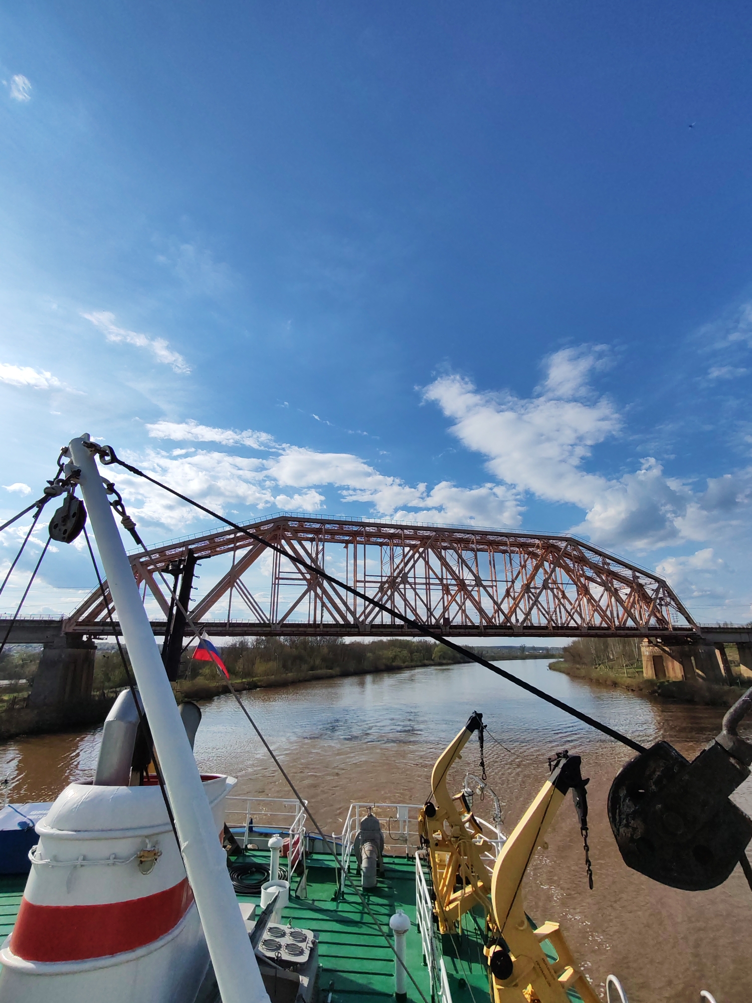A view from the deck of a vessel traversing a river, directed towards an orange truss bridge against a backdrop of a blue sky dotted with fluffy white clouds. The water in the river is brown. The deck is painted green. There are yellow cranes and a pole with ropes on the deck, and a small flag is affixed to the vessel. The location is Unknown, Unknown.