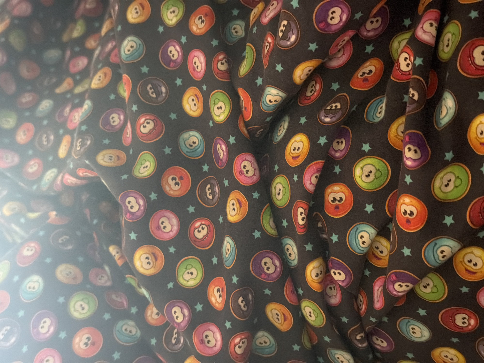 This close-up image displays a vibrant, patterned fabric draped with visible folds and shadows. The dark background is adorned with an array of colorful, cartoon-like billiard balls and small teal stars. The billiard balls come in various hues, including purple, orange, green, red, yellow, blue, black, and pink. Each ball features animated facial expressions, with wide eyes and sometimes open mouths, conveying a range of emotions from surprise to happiness or neutrality. Many of the balls are marked with numbers, predominantly '3' and '8'. A bright glare or reflection is visible on the left side of the image, suggesting an indoor setting with a nearby light source. No people are present, and no activity is taking place. The image focuses solely on the intricate details and texture of the textile pattern, offering no specific location cues for Iskar, Bulgaria.
