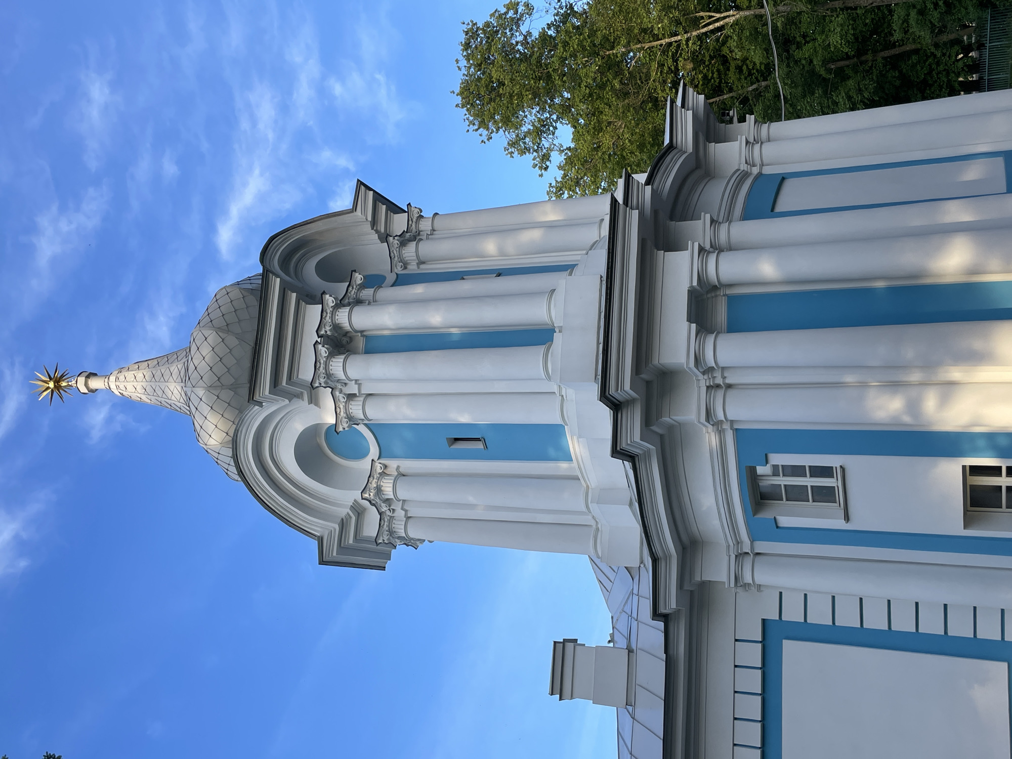 This image captures a detailed, low-angle view of one of the subsidiary bell towers of the Smolny Cathedral (Smolny Convent of the Resurrection) in Saint Petersburg, Russia. The architecture is characterized by a striking white and blue color scheme, typical of Russian Baroque.
The upper section of the tower features a silver-grey, scale-patterned dome crowned with a prominent golden multi-pointed star finial, brightly illuminated by sunlight. Below the dome, an octagonal or circular drum displays an array of tall, white Corinthian-style columns. Between these columns, recessed panels are painted a vibrant sky blue; one of these panels includes a narrow, vertical, white-framed window. Further down, the lower levels of the tower continue the white facade with blue accents, featuring a multi-paned white window on a white wall section and a partial view of another window within a blue panel.
To the left, a section of the cathedral's light grey metal roof is visible, along with a square, light grey chimney. The lighting indicates direct sunlight from the right, casting subtle shadows that highlight the architectural relief. The background consists of a clear, bright blue sky with sparse, wispy white clouds. On the right side of the frame, a dense cluster of lush green trees extends upwards, suggesting the building is set within a park or expansive grounds. No individuals are present in the scene.