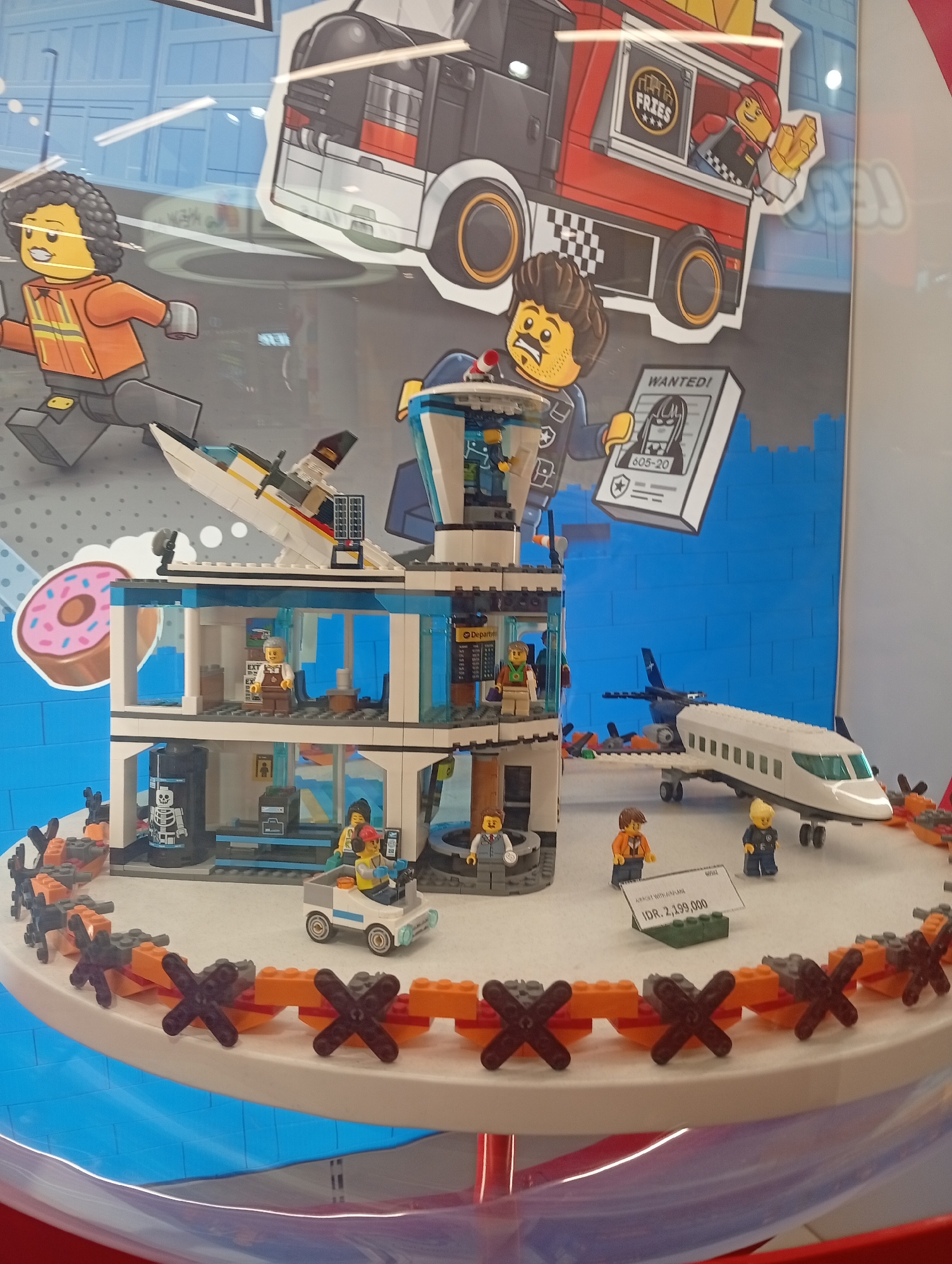 This image captures a vibrant display of Lego City sets, likely in a retail store, showcasing an airport-themed scene with additional character cutouts.
The central subject is a multi-story Lego building, resembling an airport terminal and control tower, equipped with transparent blue window elements. Several Lego minifigures are positioned within and around the building, including staff at desks, passengers, and security personnel. On the roof, a small white seaplane or boat is docked.
In front of the building, a white Lego passenger jet with a blue tail is parked, along with a small white ground vehicle driven by a minifigure. The entire Lego setup rests on a round, white display table, bordered by an orange and black fence-like structure made of Lego bricks.
The backdrop features large, colorful cardboard cutouts of Lego minifigures and vehicles, creating a dynamic, action-oriented scene. These include a running minifigure with curly hair and an orange jacket, a minifigure holding a "WANTED!" poster, and a red food truck with "FRIES" written on it. A large pink donut graphic is also visible. The background also features a light blue brick pattern.
The display is enclosed behind a reflective glass or plastic barrier, evident from the reflections across the image. A white price tag on a green Lego brick base is visible in the foreground, displaying "IDR. 2.199.000," indicating the price in Indonesian Rupiah. The setting suggests an indoor environment, such as a toy store in Denpasar, Indonesia.