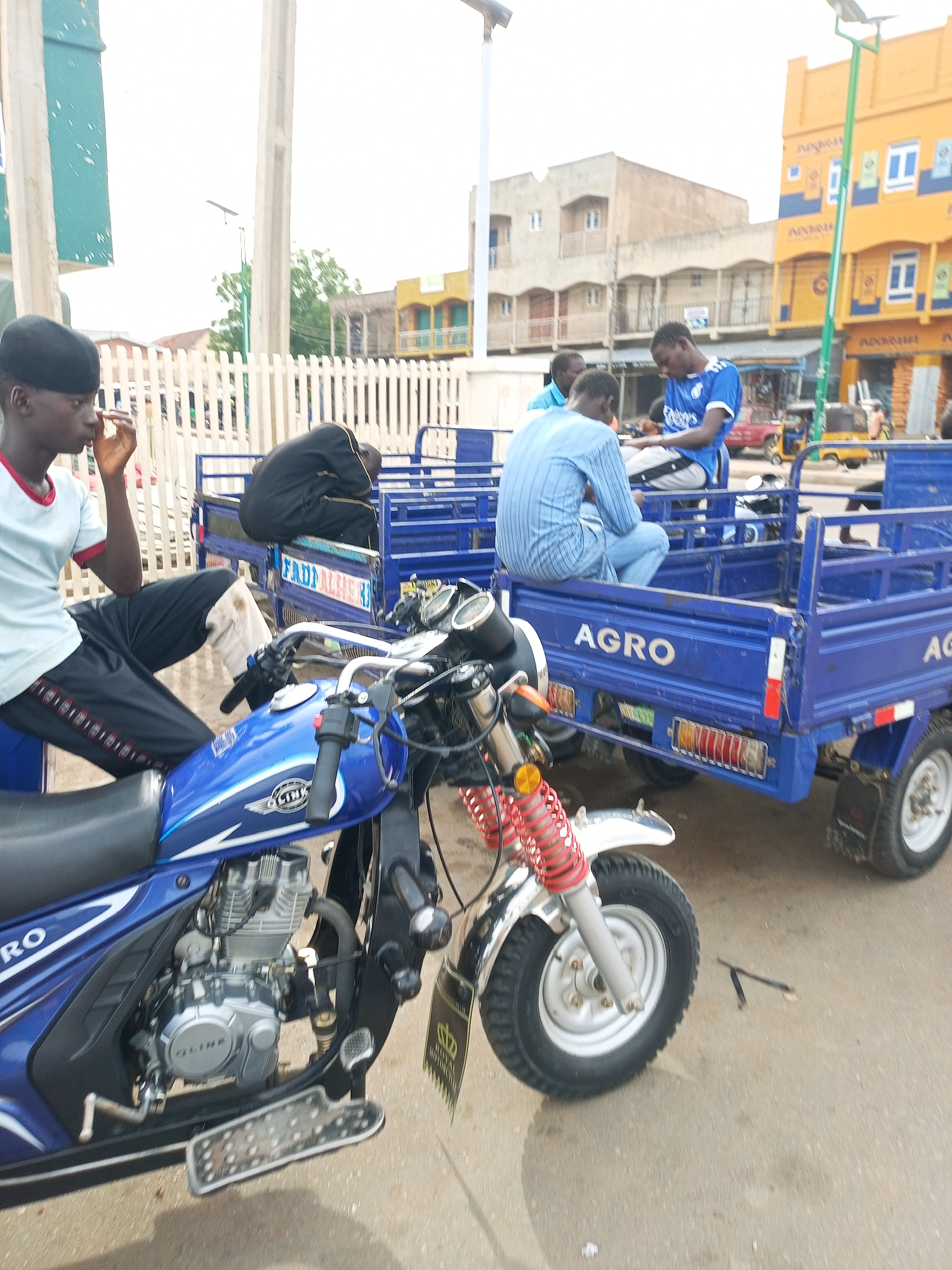 In Arawa, Nigeria, a blue motorcycle trike is parked on a paved surface. The motorcycle features a silver engine, black seating, and silver handlebars. A person wearing a light blue t-shirt with red trim and black pants sits on the motorcycle. Next to the motorcycle, a blue cargo trike is visible, with "AGRO" printed on its side. Three individuals are seated in the cargo trike. One man wears a blue and white striped shirt and jeans, another wears a blue t-shirt, and the third is dressed in black. Behind them, buildings with yellow and light brown facades are present, with an overcast sky overhead.