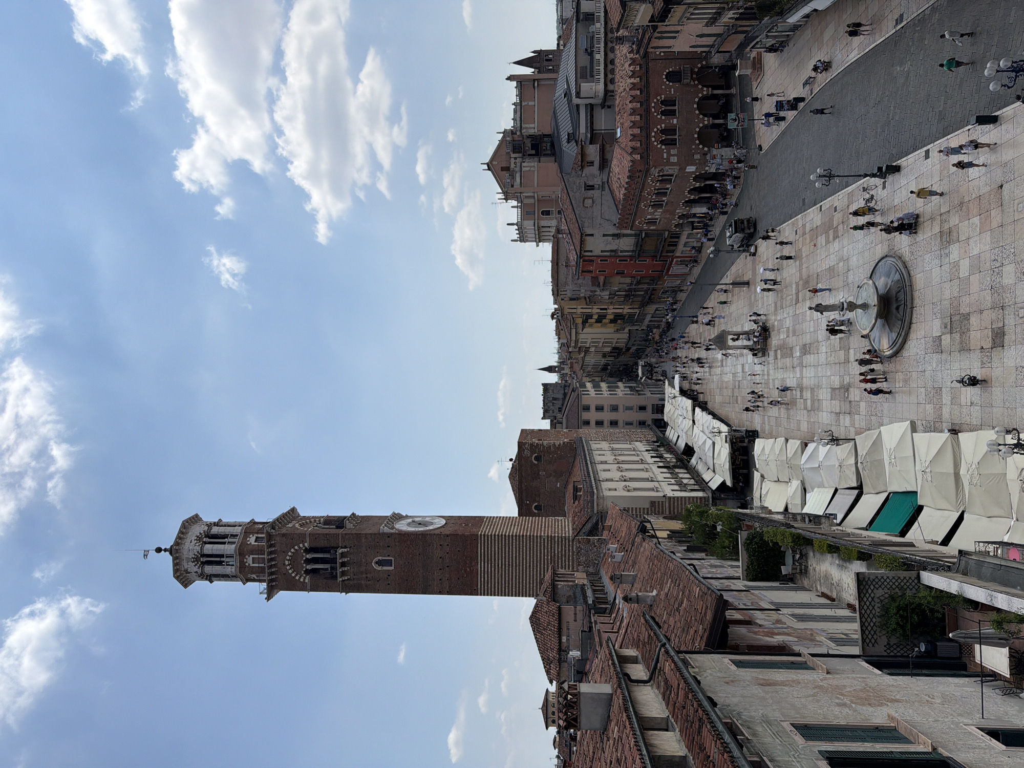 This is a high-angle, eye-level, full shot of Piazza Bra in Verona, Italy, on a bright, sunny day with scattered clouds. The iconic Torre dei Lamberti, a tall brick clock tower, dominates the left side of the frame. Its distinctive white stone upper levels and Romanesque architecture are clearly visible, along with a large clock face on its side.
The piazza itself is paved with a light-colored, patterned stone. A long row of white market stalls or awnings stretches along the right side of the square, casting shadows on the ground. People are dispersed throughout the piazza, some walking, others congregating near a fountain that features a statue. A few bicycles are parked near the foreground.
The buildings lining the piazza are a mix of architectural styles, with tiled terracotta roofs and facades in various colors, including cream, red, and ochre. The architecture suggests a historic European city. In the background, more buildings are visible, some with arched windows and intricate details. A white truck is present on the street bordering the piazza. The scene depicts a lively, urban public space during daylight hours.