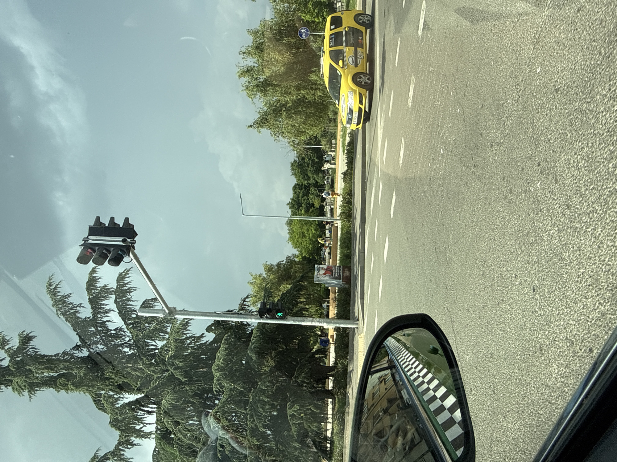 A yellow taxi is turning right at an intersection in Burgas, Bulgaria, under a cloudy sky. The taxi has a checkered stripe on its side and a "6+1" sticker. In the foreground, the side mirror of the vehicle from which the photo is taken reflects a checkered pattern, likely on the car itself. Across the street, there are trees and people walking on a sidewalk. A traffic light shows a green signal for pedestrians. Another traffic light, visible on a tall pole, shows red. A billboard with an advertisement is also present. The road surface is asphalt with white lane markings.