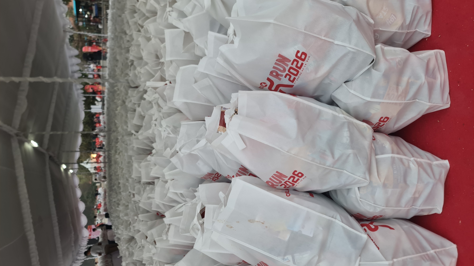 The image depicts a vast quantity of white reusable tote bags, densely packed on a red surface, likely a table or the ground. Many of these bags are adorned with a red graphic and the text "RUN 2026 SERIES OF CK RUN." In the background, beneath a white tent structure, numerous people in casual attire, many wearing red, are visible. They appear to be participating in or organizing an event, possibly a race given the "RUN" text on the bags. The overall scene suggests a pre-event distribution or preparation for a large-scale running event. The location is identified as Denpasar, Indonesia.