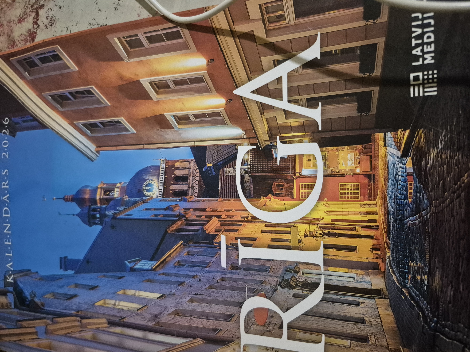 The image is a calendar or promotional material featuring a nighttime view of a cobblestone street in Riga, Latvia. The street is lined with historic buildings, some of which are illuminated. In the foreground, large white letters spell out "RIGA." In the background, a tall clock tower with a dome is visible. The sky is dark blue, suggesting it is evening or night. At the bottom right of the image, a logo with the text "LATVIJAS MEDIJI" is present.