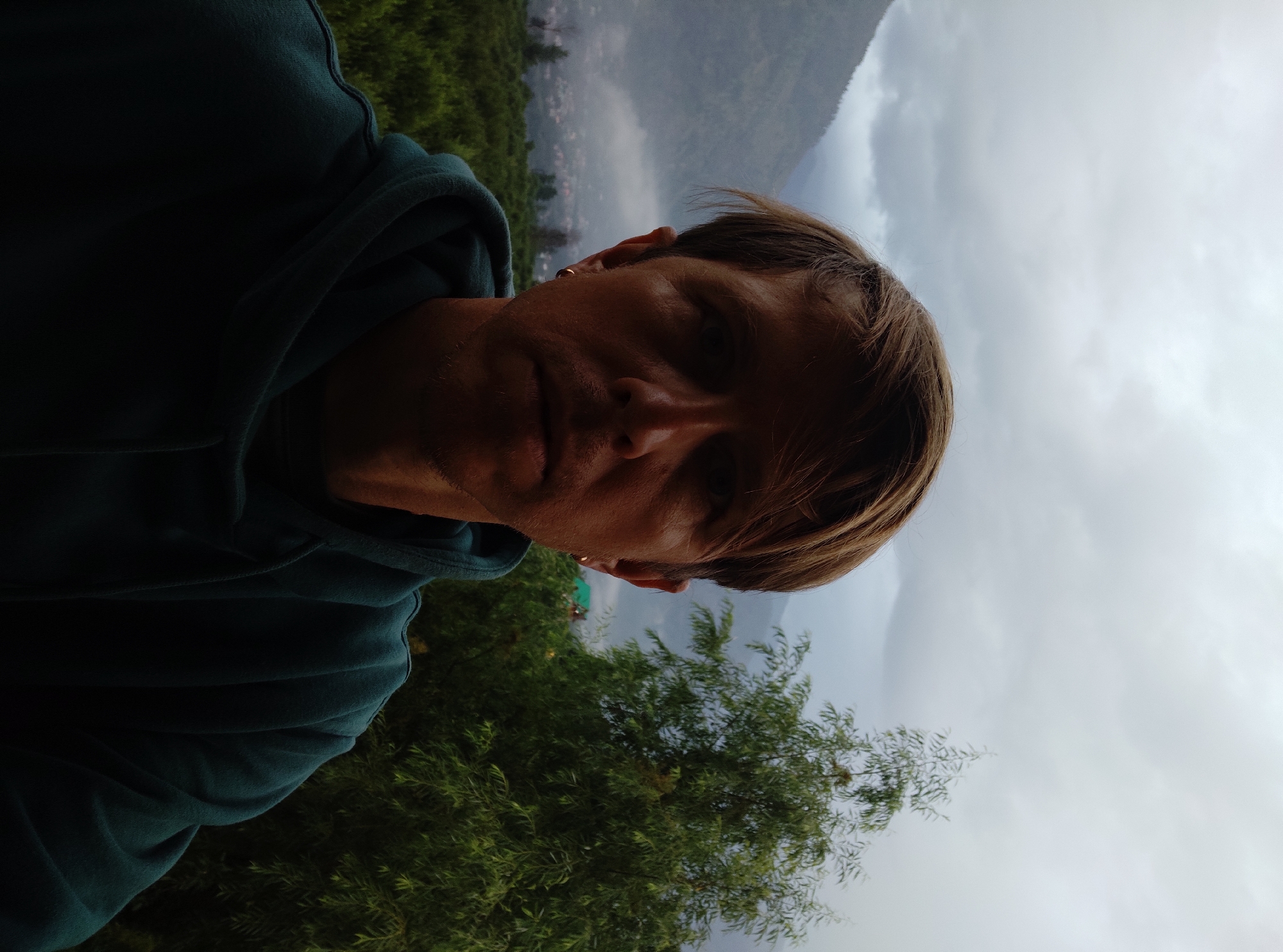 A close-up, front-facing selfie features a light-skinned male with light brown hair and blue eyes against a cloudy sky. He is wearing a dark teal hooded sweatshirt. In the background, a dense green forest extends to a valley with a village, and further back, misty mountains rise under a grey, overcast sky. The image is taken outdoors, likely in a mountainous region. The man is looking directly at the camera, and his expression is neutral. He has a small gold hoop earring in his left ear.