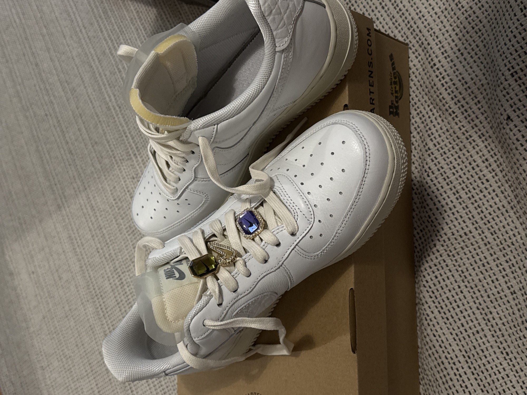 Two white low-top sneakers are displayed in an indoor, still-life setting, likely during the evening or night under artificial light, in Hainburg an der Donau, Austria. The shoes are placed on a brown cardboard box, partially visible.
The shoe in the foreground, angled towards the viewer, features two decorative rings adorning its off-white laces. One ring is rectangular with a prominent blue gemstone, while the other is an oval-shaped green/gold gemstone, positioned closer to the shoe's tongue. A "NIKE" logo is visible on the cream-colored tongue of this shoe. The shoe has small perforations on its toe box.
The second sneaker is positioned behind and slightly to the left of the first, with its sole facing the viewer. Both shoes appear to be made of white leather with light-colored soles and textured accents around the ankle collar.
The cardboard box beneath the shoes displays branding for "Dr. Martens," including "AIR WAIR" and "MARTENS.COM" text. The background consists of a light-colored, textured fabric, possibly a carpet or blanket, providing a soft contrast to the footwear. There are no people or discernible activities taking place beyond the arrangement of the shoes for display.