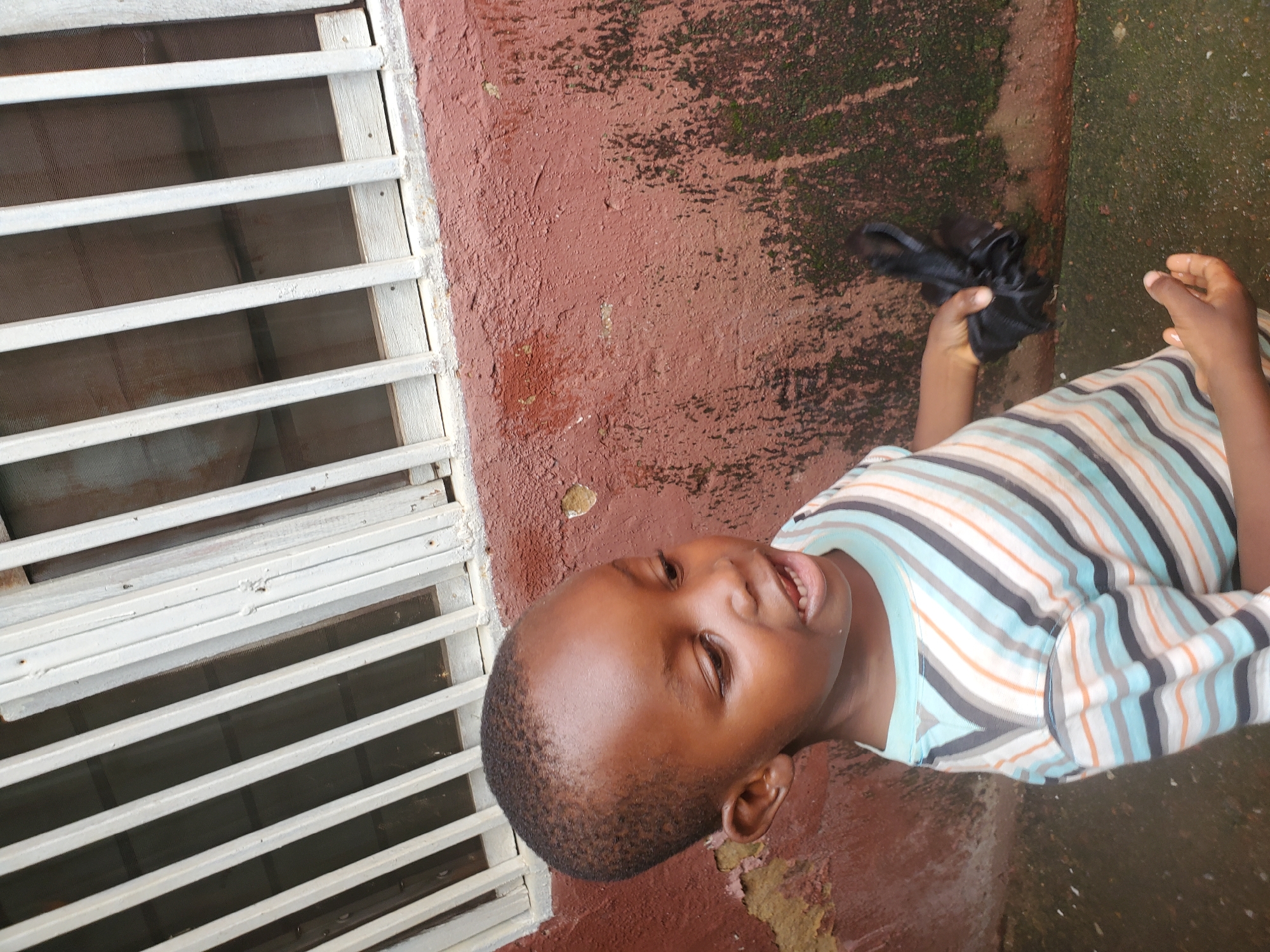 A young Black boy stands outdoors in front of a red, textured wall with a window grate above. He is wearing a striped t-shirt in shades of blue, gray, and orange. In his right hand, he holds a crumpled black object. His left hand is visible at his side. The wall shows signs of wear and some green moss growth. The ground appears to be wet and dark, possibly asphalt or concrete. The lighting suggests an overcast day.