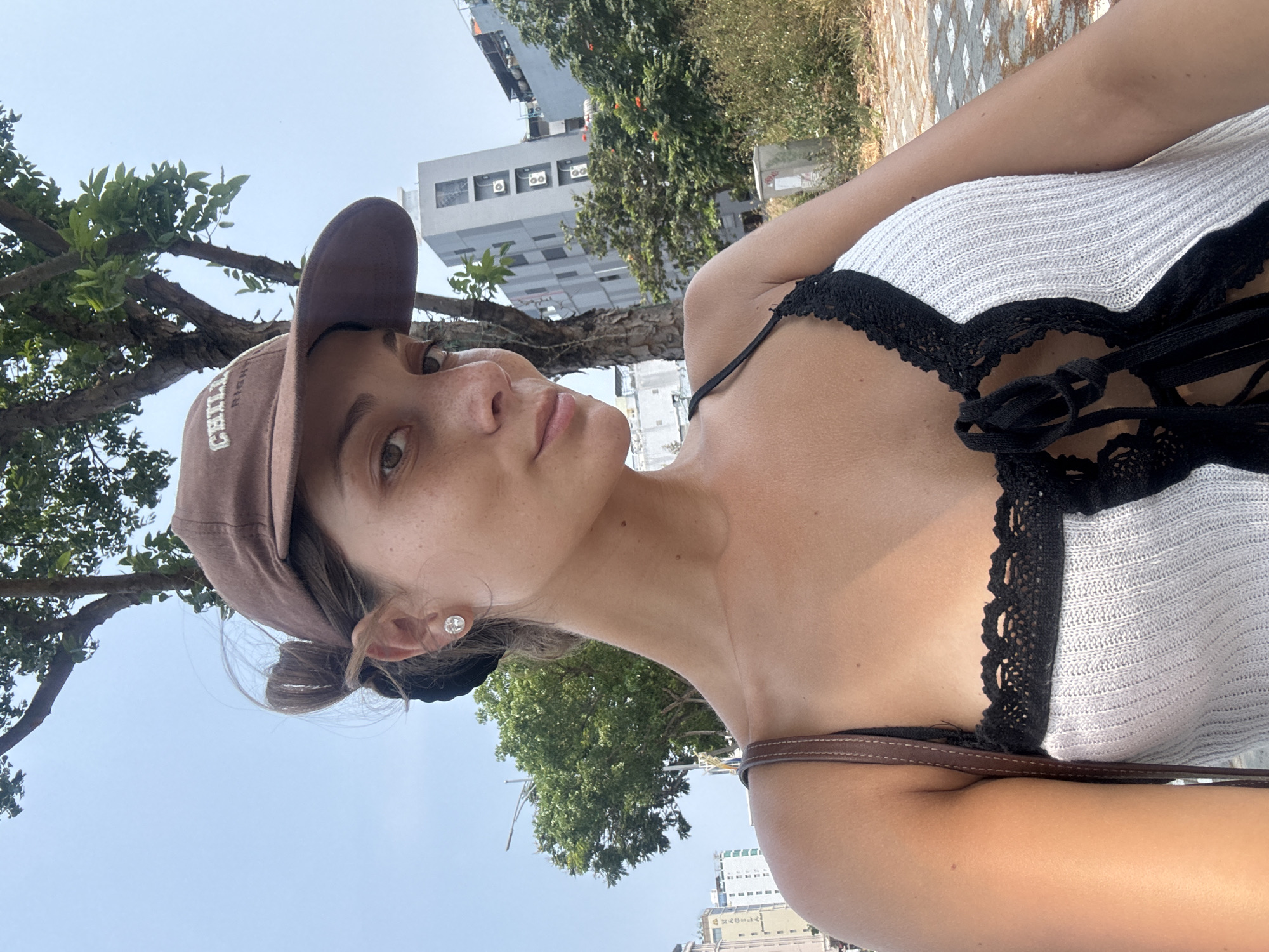 A woman is posing for a selfie outdoors during the day. She is wearing a brown baseball cap, a white top with black lace details, and a small earring. She is situated in an urban environment, likely in Đà Nẵng, Vietnam. There are trees, buildings, and a clear sky visible in the background. The perspective is upward, with the camera held at a low angle.