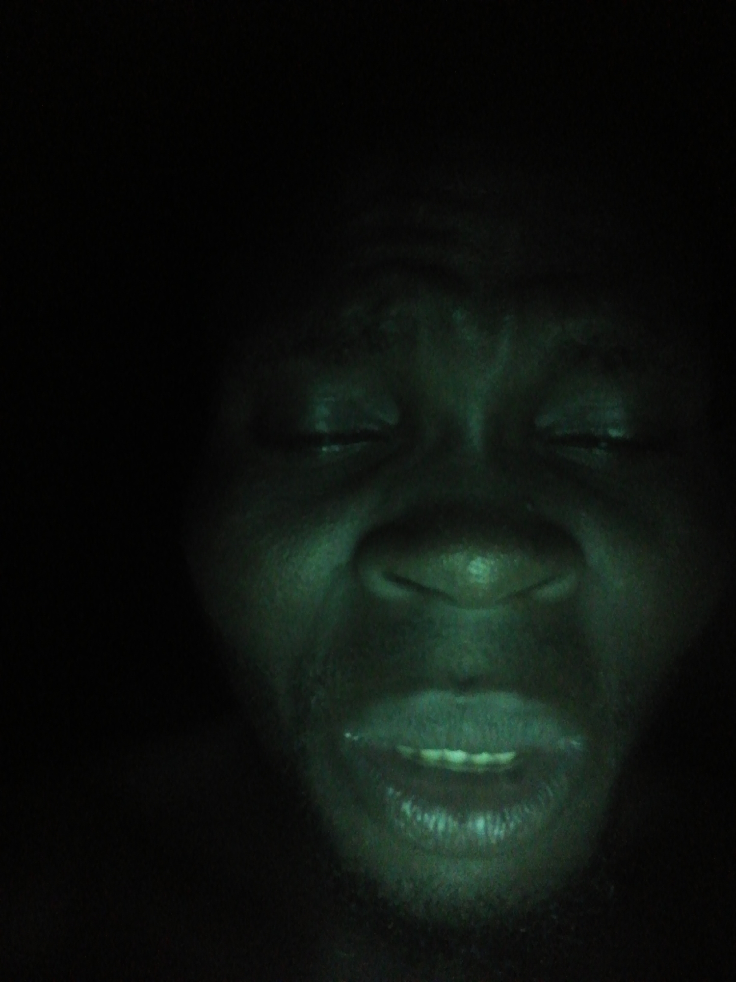 A close-up, low-light image of a person's face. The person has dark skin and closed eyes. Their mouth is slightly open, revealing some teeth. The surrounding environment is dark and indistinct. There are no additional details to identify a location.