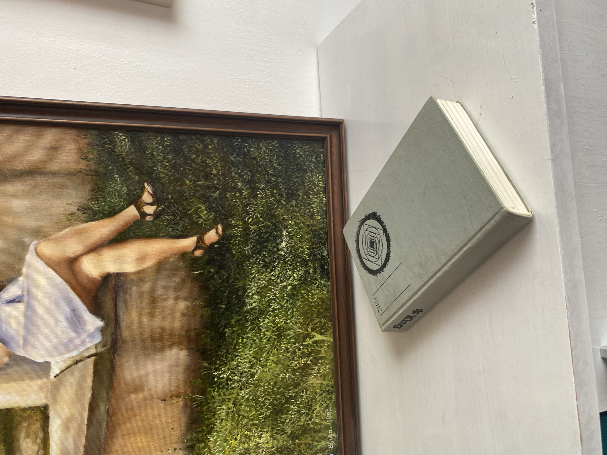 An oil painting depicting a woman's legs and torso, wearing a white dress and sandals, is leaning on a stone ledge with a grassy background. The painting is framed and placed on a white surface next to a light grey hardcover book. The book's spine is visible, showing the word "King" and a circular emblem with geometric patterns. The title "Maj" is also present on the book's cover, along with a smaller emblem. The scene appears to be indoors, on a white shelf or table, with a white wall in the background. The painting is signed "Bljna, 2020" in the lower left corner. The location is Bílina, Czechia.